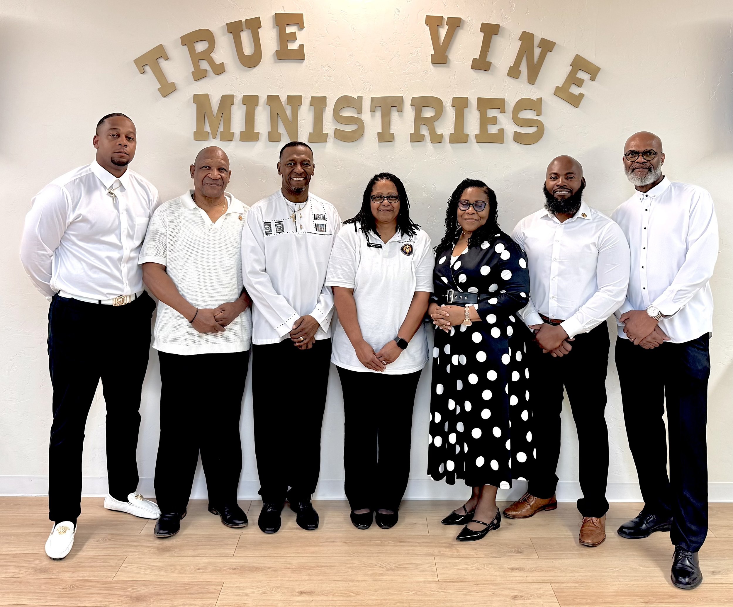 Group of seven people standing in front of a wall with large beige letters that spell "TRUE YIN MINISTRIES." The group is dressed in semi-formal attire, smiling and posing for the photo.