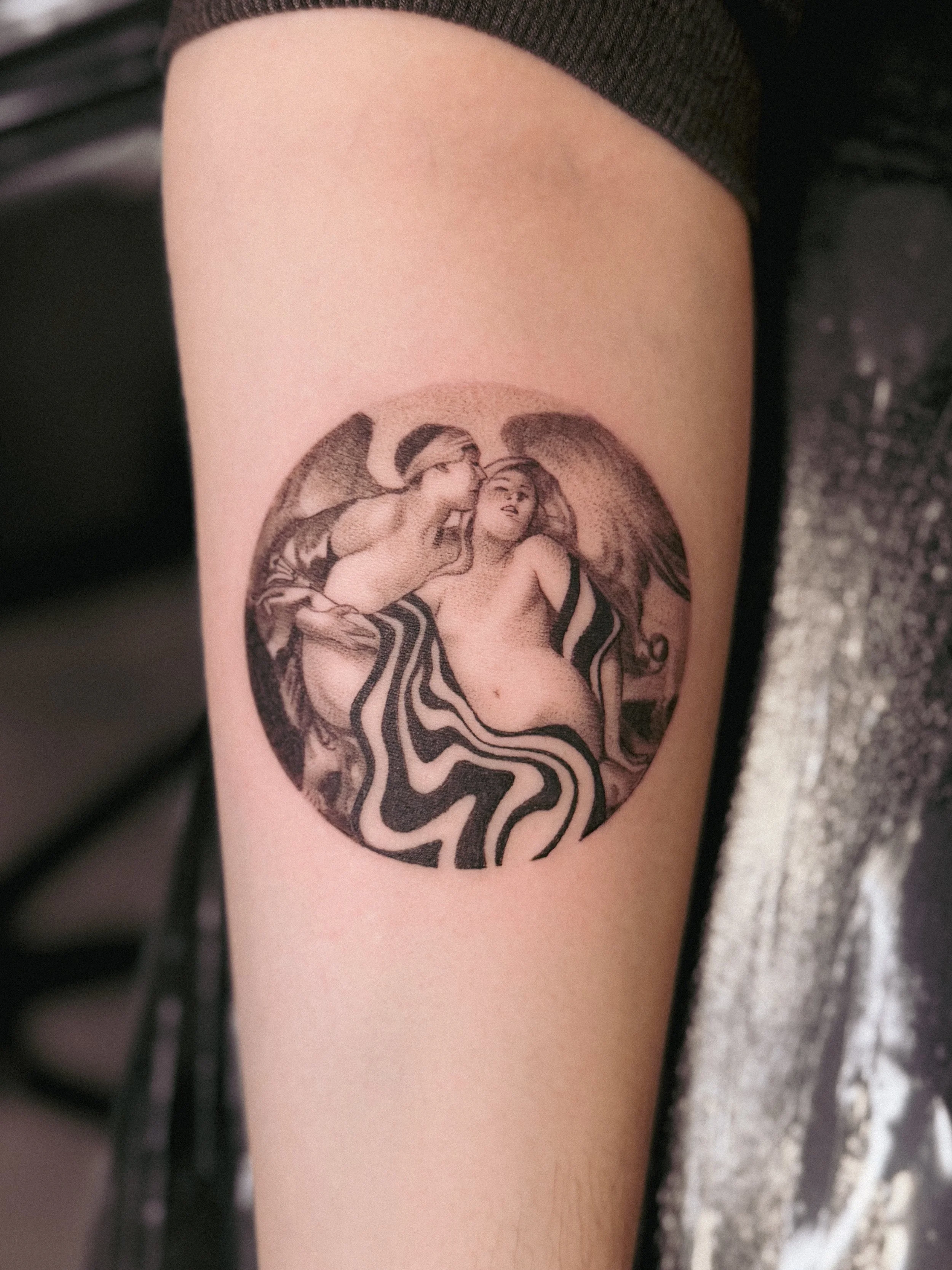 Tattoo of two women with wings, sitting close together, with striped fabric below them, on a person's inner arm.