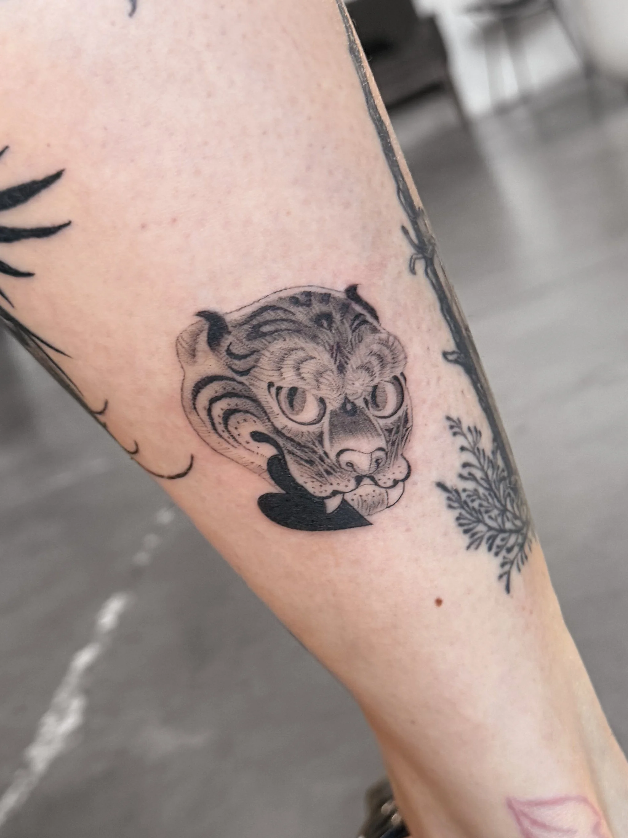 Close-up of a tattoo on a person's arm, featuring a stylized lion's face with intricate lines and shading, and additional part of the tattoo visible on the right side of the image. tiger asian tattoo