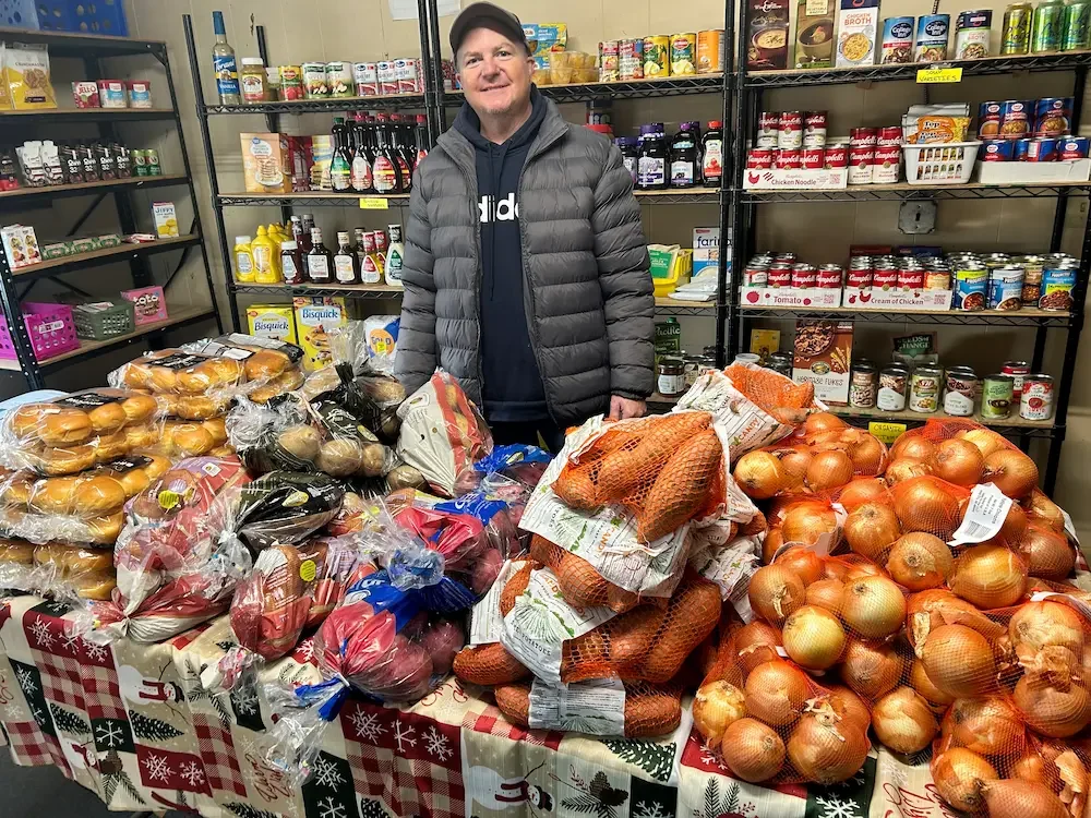 A man standing behind a table filled with onions, garlic, and bread in a grocery store aisle.