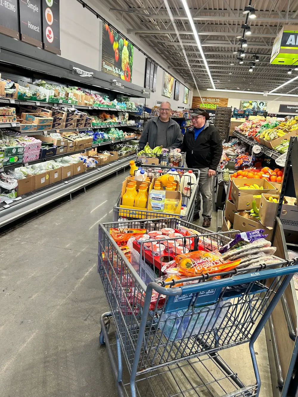 Two men shopping for groceries in a supermarket, standing at a shopping cart filled with bottled drinks, packaged foods, and produce. The store has shelves of vegetables, fruits, and other items, with bright overhead lighting and large digital menu s