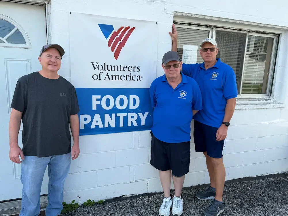 Three men standing outside next to a 'Volunteers of America Food Pantry' sign, with one man wearing a gray T-shirt and two men in blue shirts, one with a cap and sunglasses.