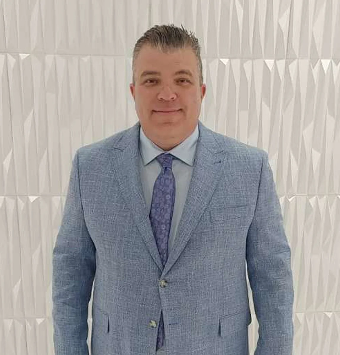 A man in a light blue suit with a purple tie, standing in front of a white, textured wall.