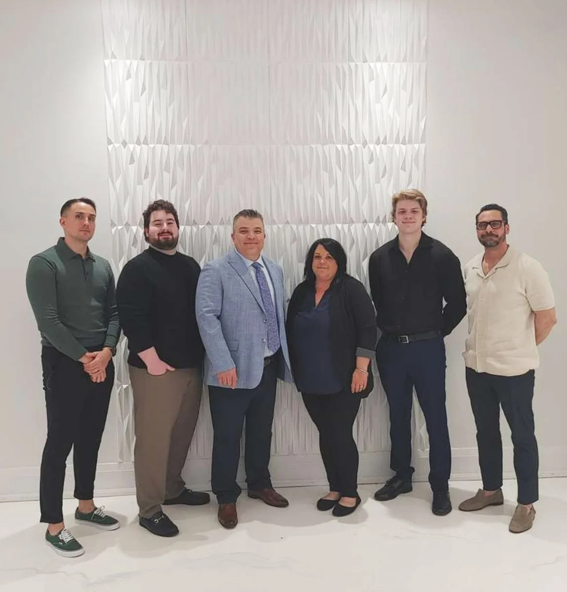 Group of six people standing indoors in front of a textured white wall.