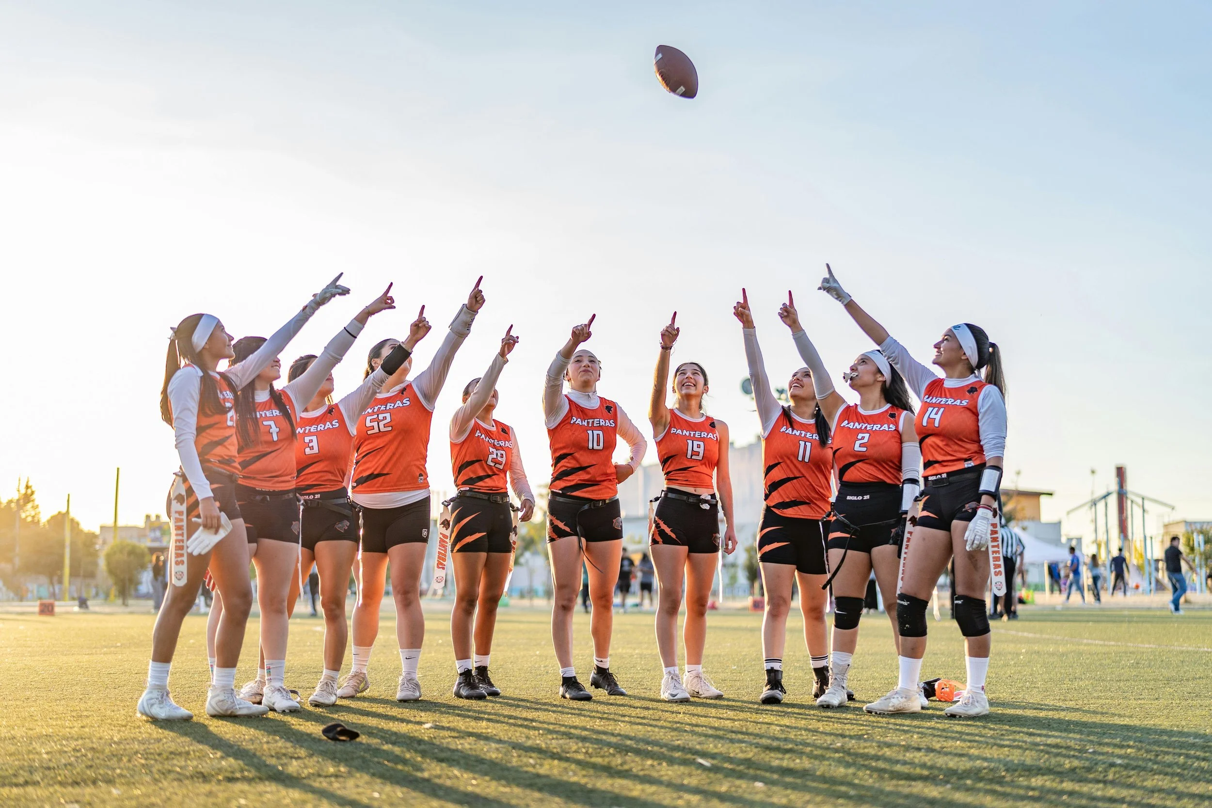 A group of female flag football players wearing orange and black uniforms, standing together on a field during sunset, some pointing at a football in the sky.