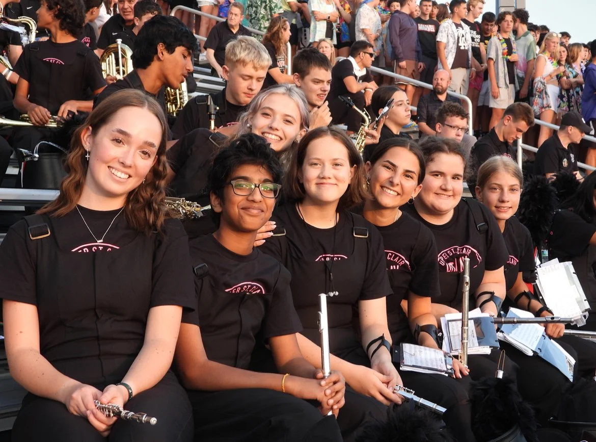 Group of young women in black uniforms with musical instruments, sitting and smiling at a band performance or event, with a crowd of people in the background.