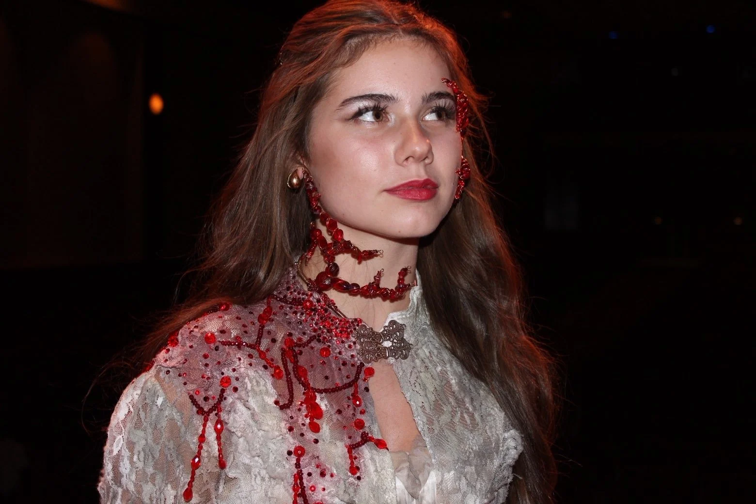 A woman with long brown hair wearing a lace dress and red jewelry, including necklaces, earrings, and earrings, standing against a dark background.