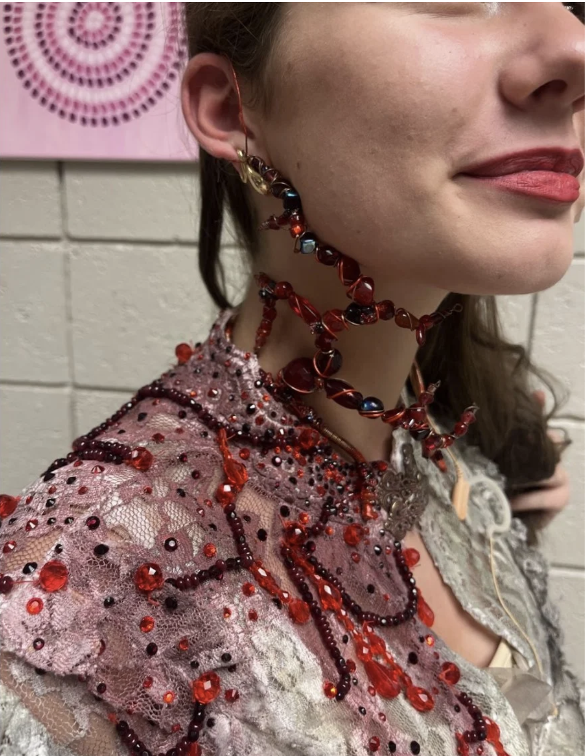 A woman wearing a pink lace dress adorned with red and black beads and jewelry. She has multiple necklaces and earrings made of red and black beads, with some metallic accents. Her lips are painted red, and she is standing against a brick wall backgr