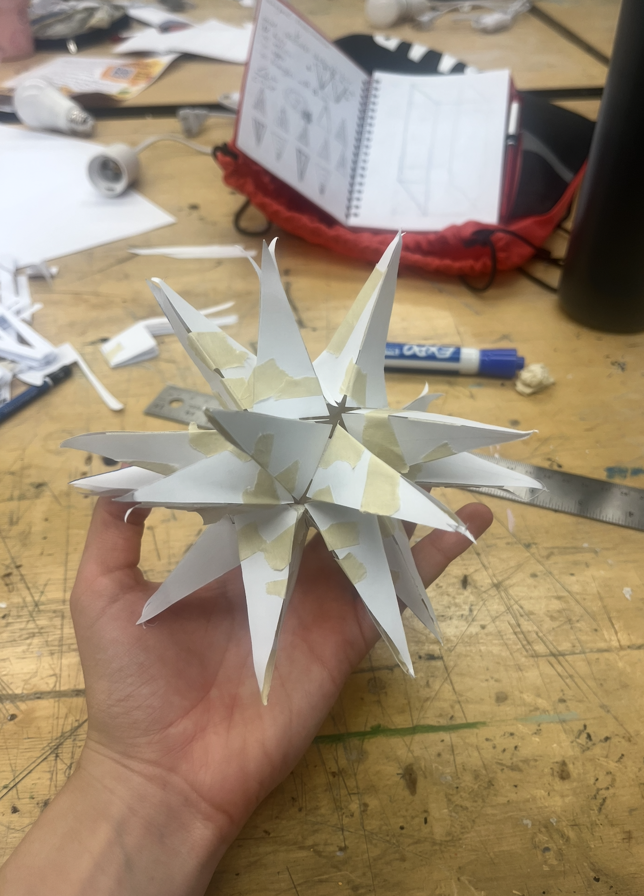 A hand holding a white paper star ornament with multiple pointed spikes, taped with masking tape. The background shows a cluttered worktable with craft supplies, a marker, ruler, and a spiral notebook containing diagrams.