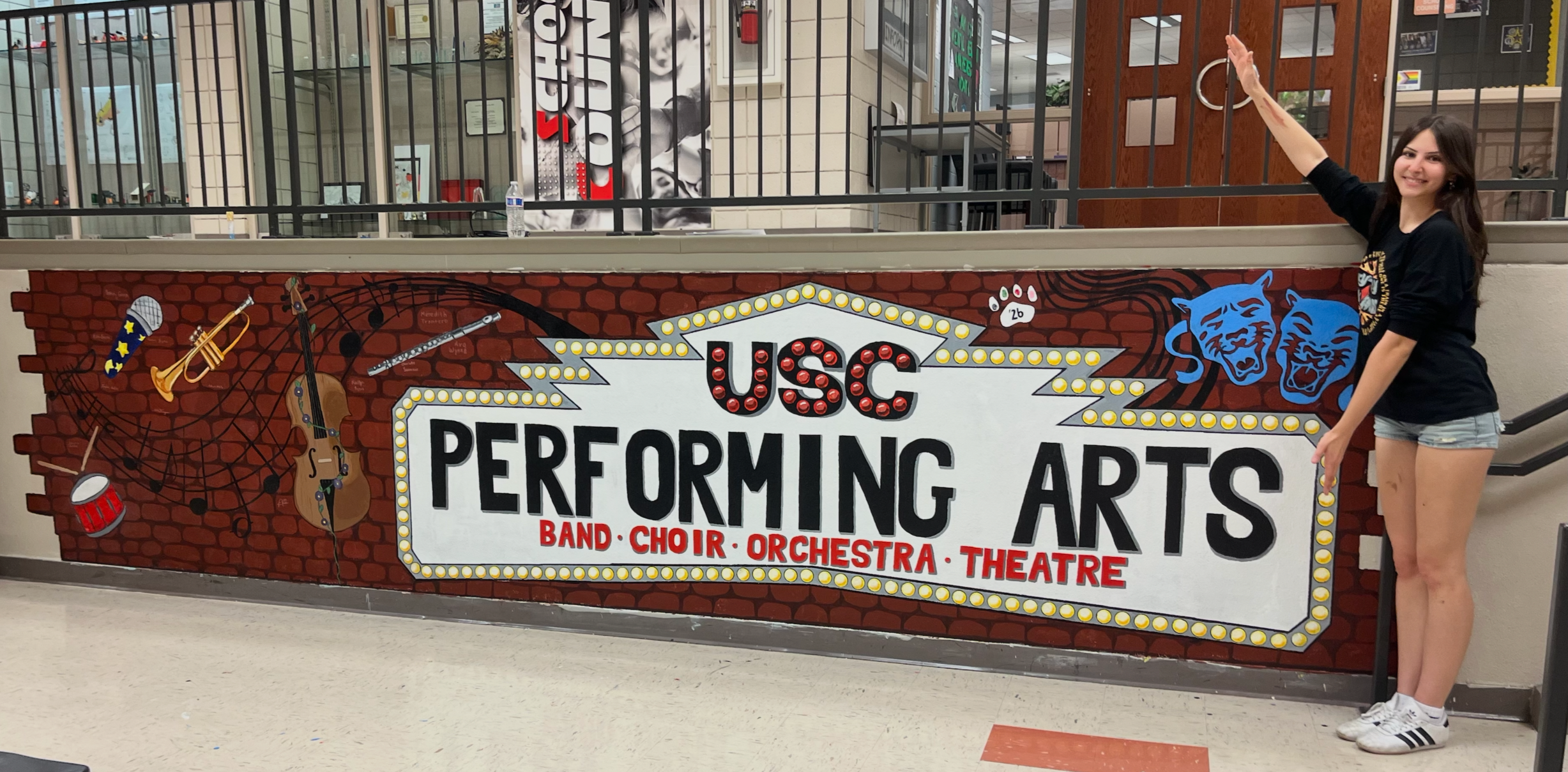 A girl with long dark hair wearing a black t-shirt and denim shorts pointing and smiling next to a colorful sign for the US Corps Performing Arts, which includes symbols of a microphone, trumpet, guitar, drum, and two blue panther heads.