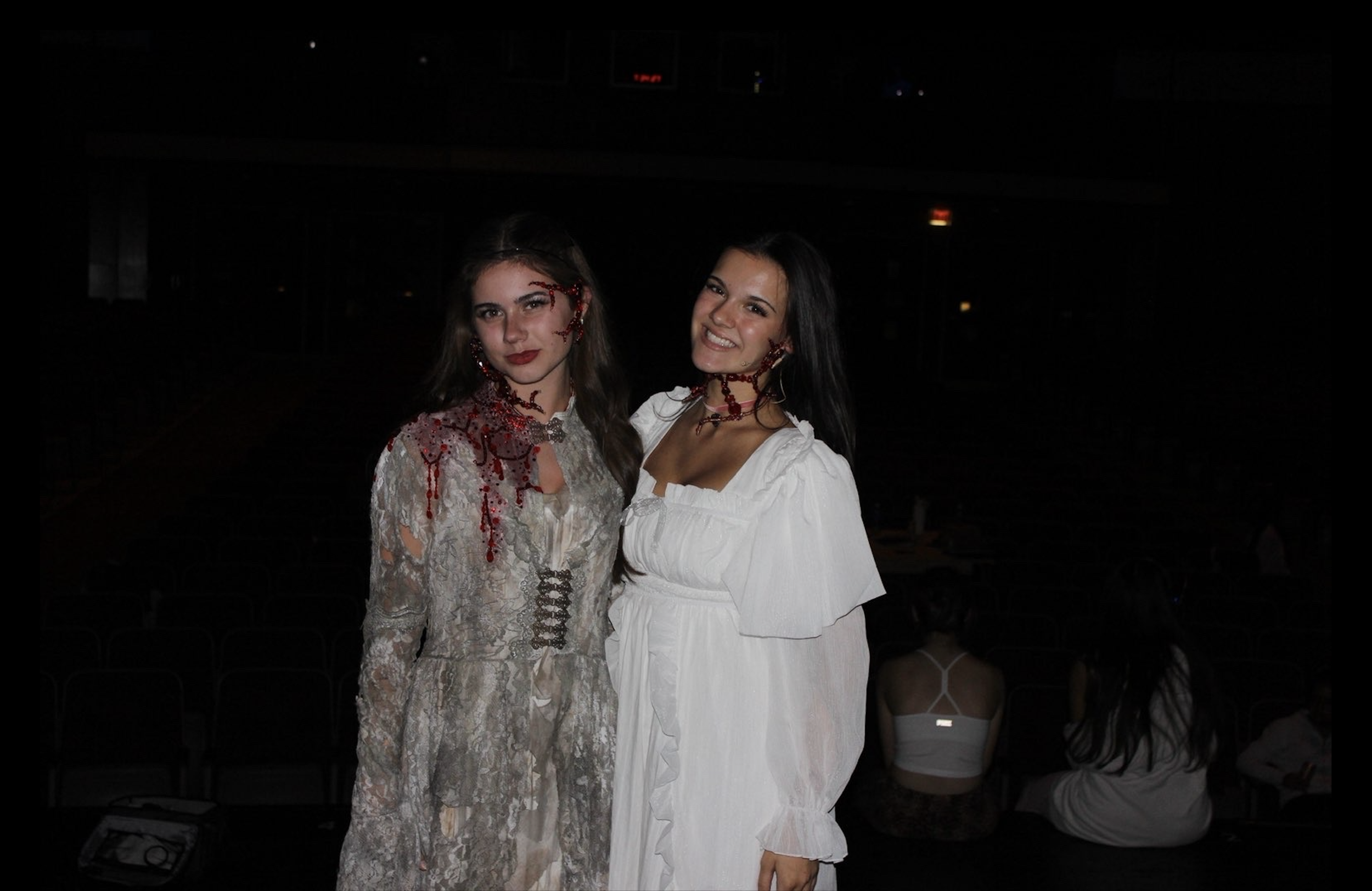 Two women are standing close together, smiling at the camera in a dark indoor setting, with other people faintly visible in the background.