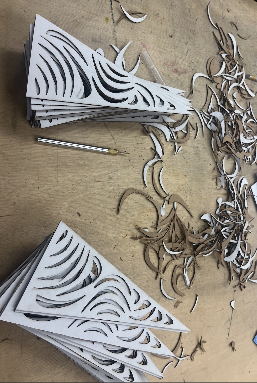 Laser-cut paper or cardboard pieces in various shapes, possibly preparing for an art or craft project, with a cutting tool on a wooden work surface.