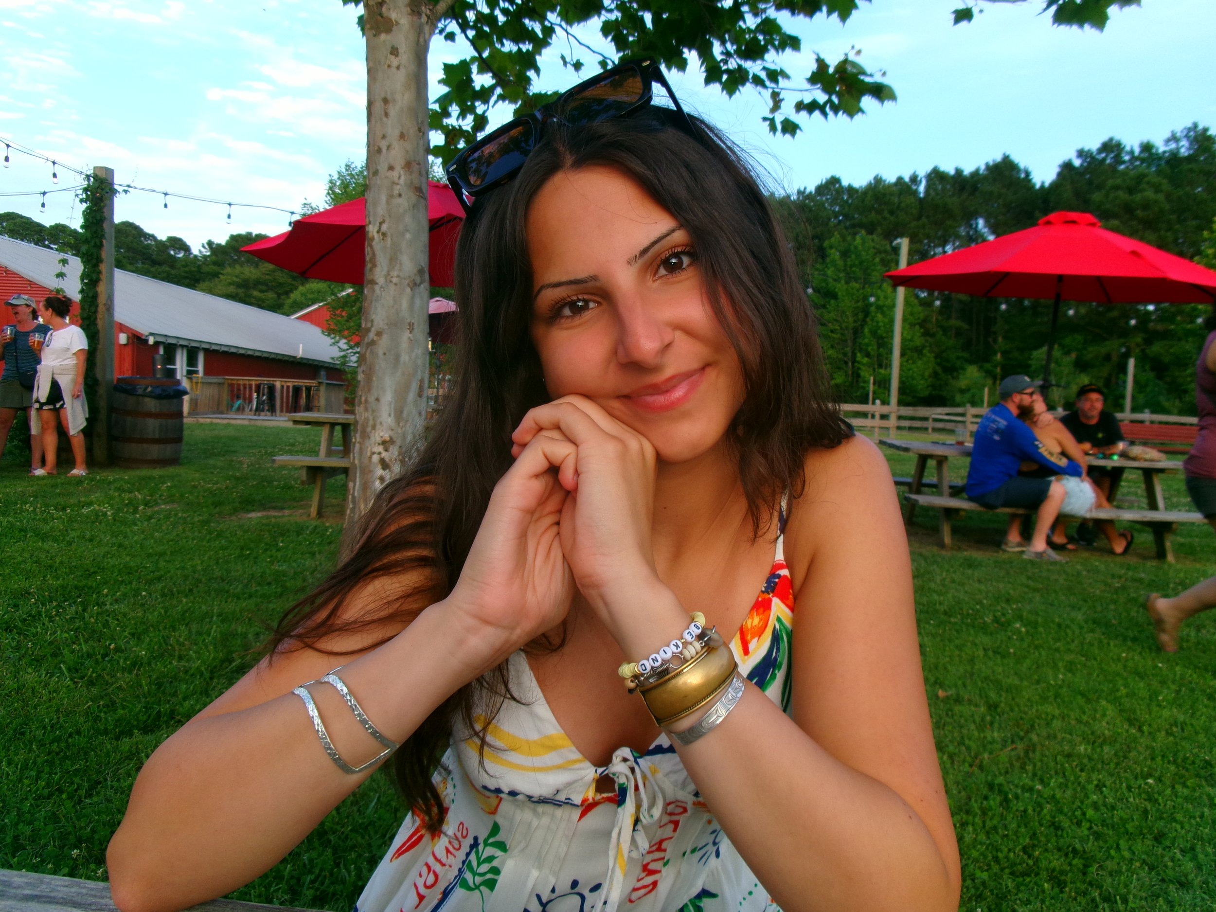 A young woman with long dark hair, wearing a colorful dress, smiling, sitting outside at a park or outdoor area with red umbrellas and other people in the background.