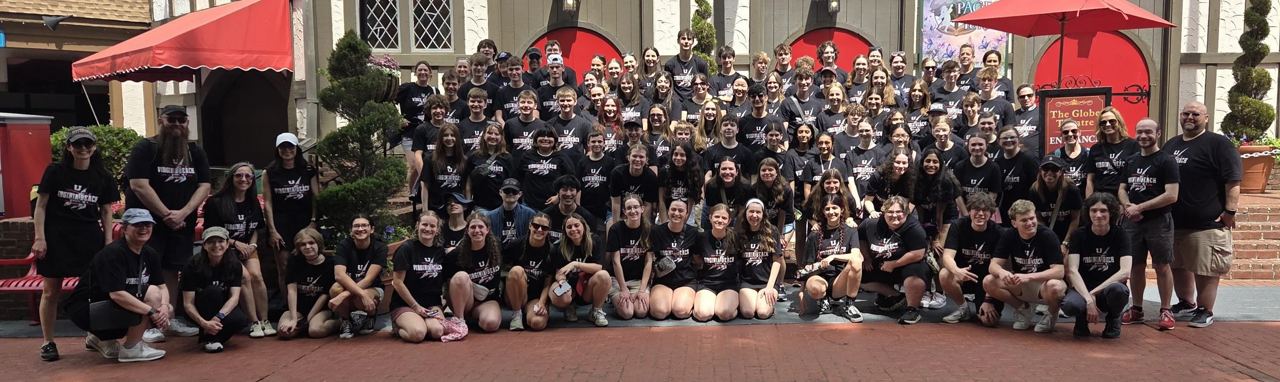 A large group of young people and a few adults posing together outside on a brick-paved area with a backdrop of a building and greenery, all wearing black T-shirts with a logo, during daytime.