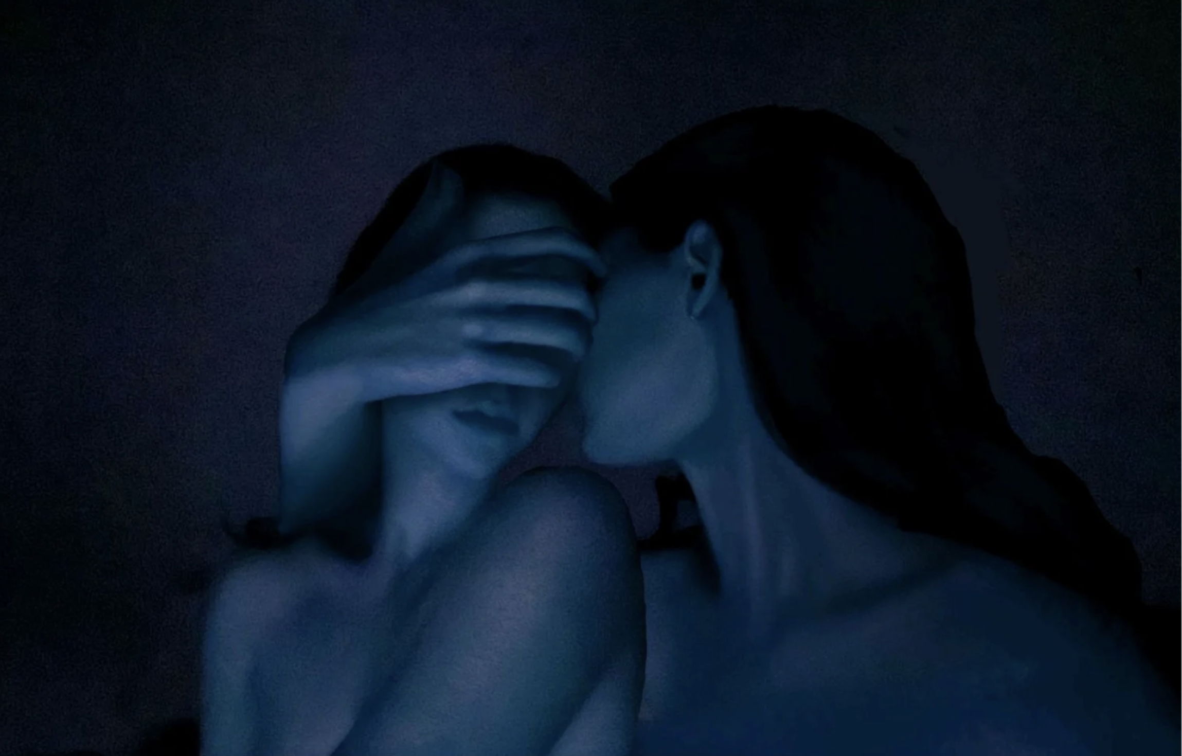 A person with dark hair and light skin is shown in a dark environment, holding their hand over their face, with fingers touching the forehead and mask.