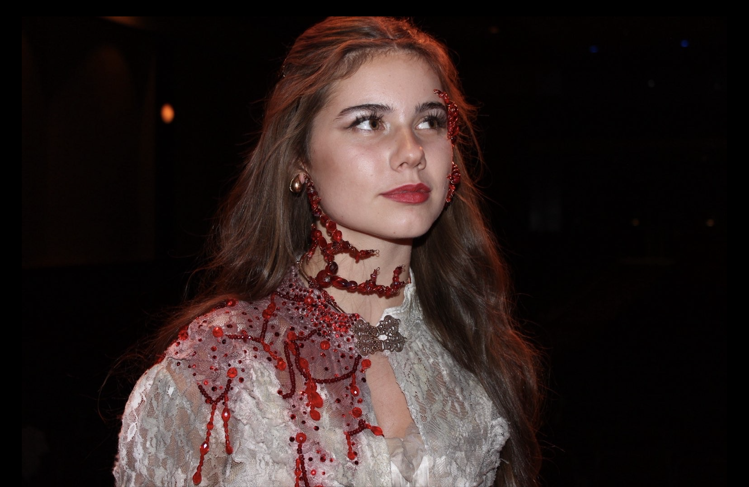 A woman with long brown hair wearing red jewelry and a lace dress with red embroidery.