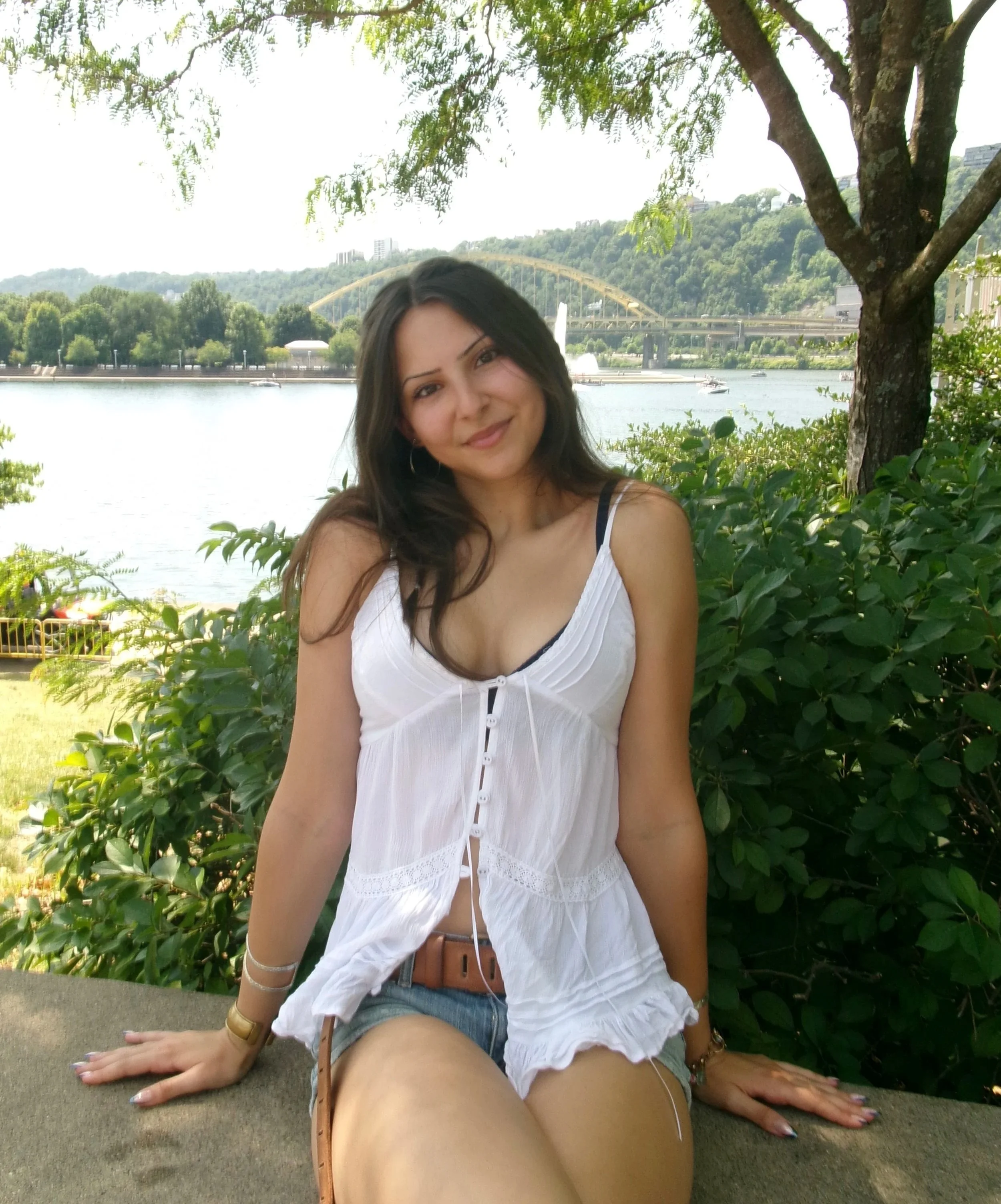 A woman sitting outdoors near a river, with green foliage and a bridge in the background. She is wearing a white top with a black strap and denim shorts, and appears to be smiling.
