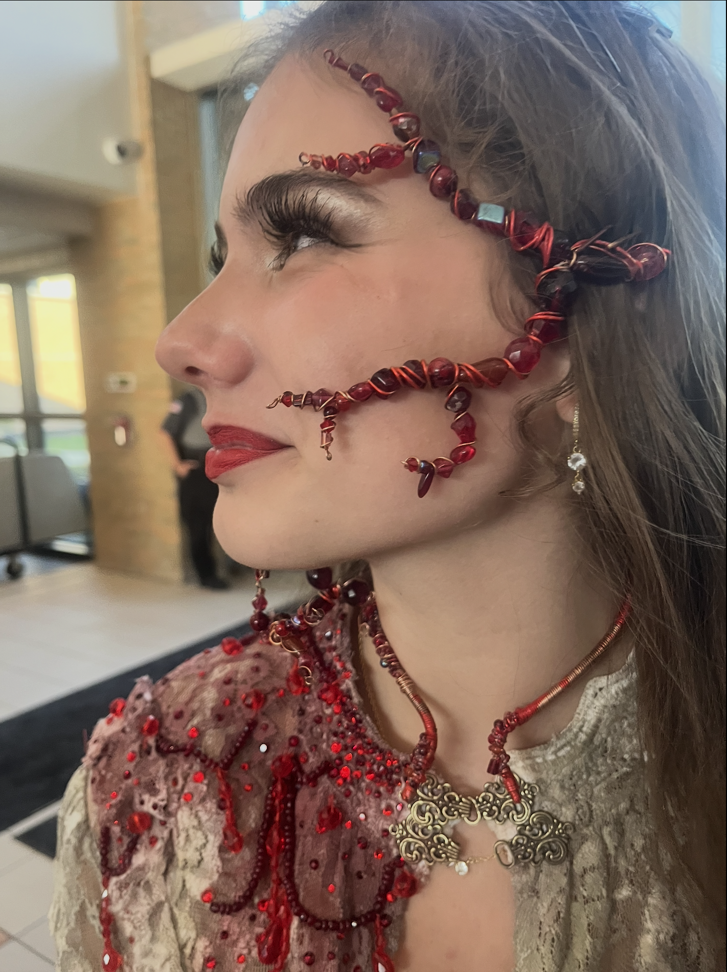 Close-up of a woman with elaborate red jewelry, including a beaded face chain, earrings, necklace, and hair accessories, wearing a lace dress with red embellishments, in an indoor setting.