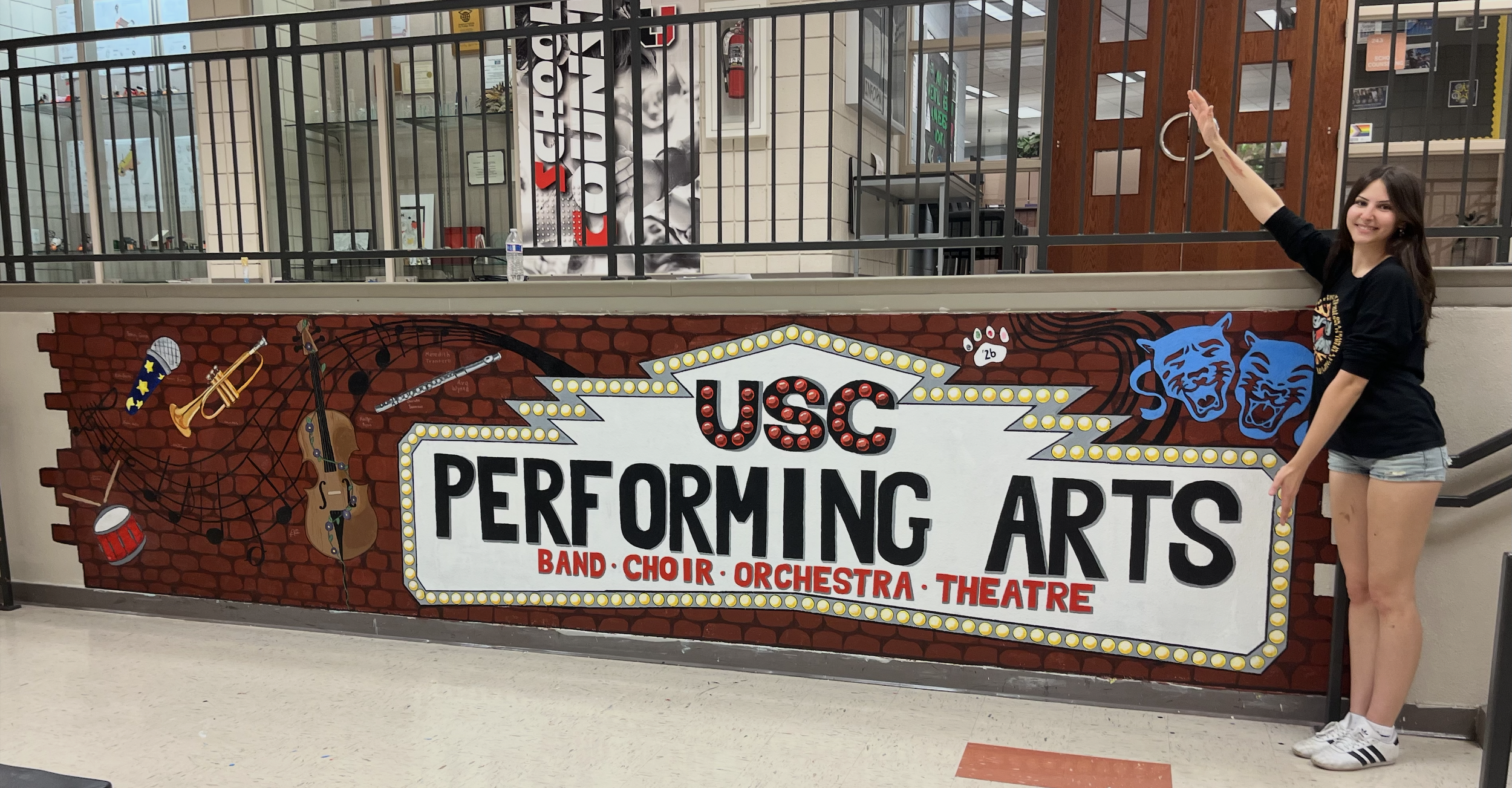 Young woman in black shirt and denim shorts pointing to a sign for a performing arts center with a brick wall background decorated with musical instruments and blue panther faces.