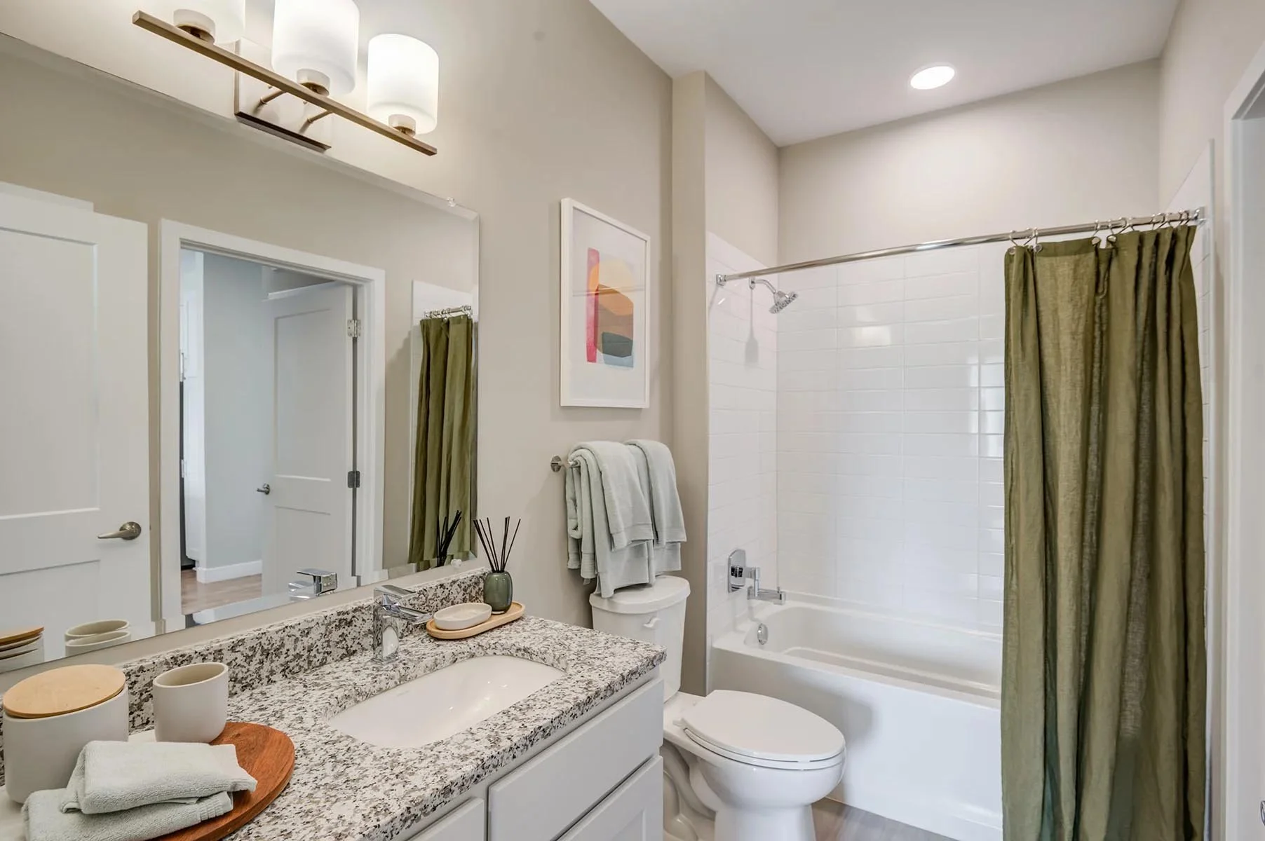 Bathroom with a granite countertop, a large mirror, and vanity items. Towels hanging on a rack, abstract artwork on the wall, a bathtub with a shower and curtain, and a toilet.
