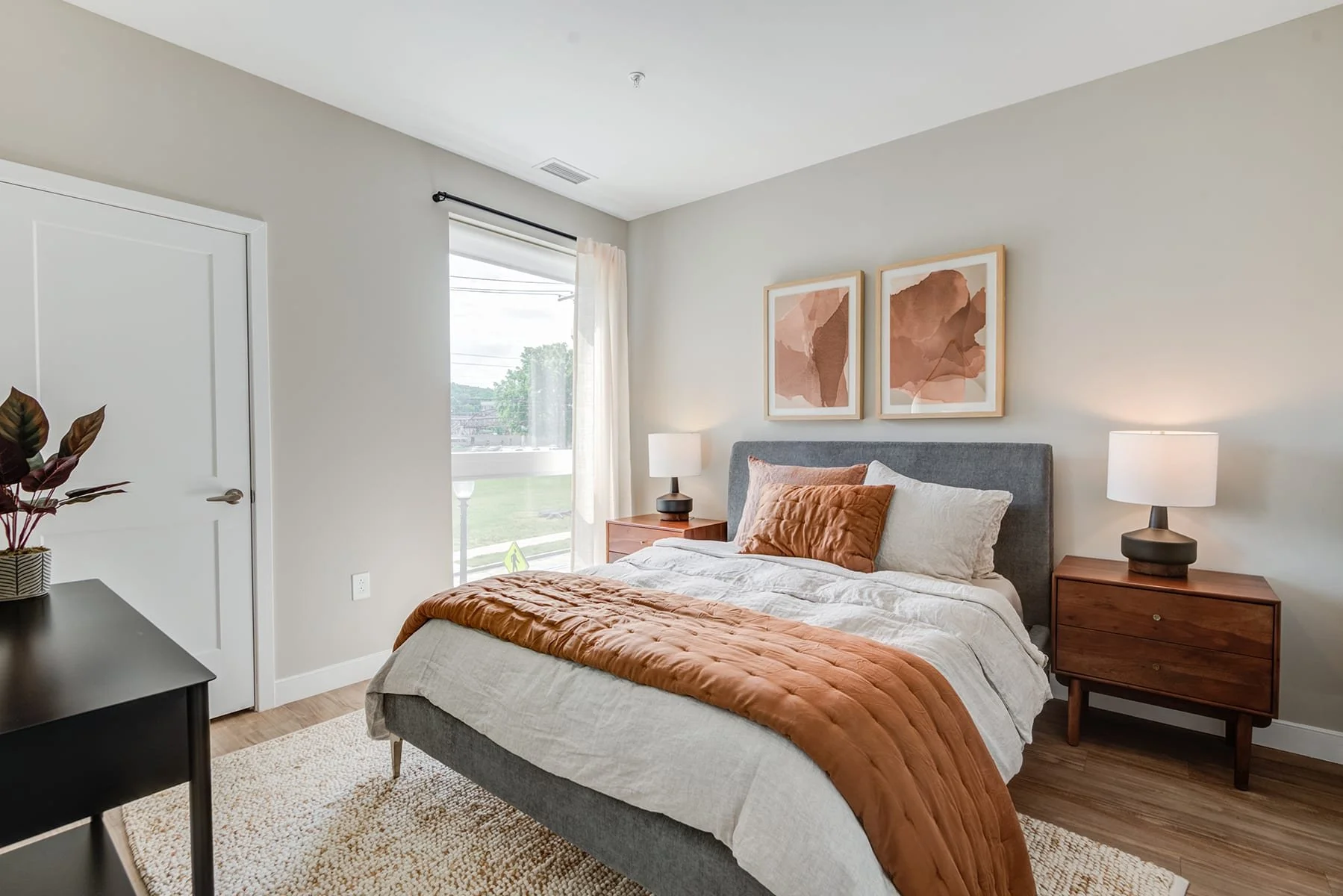 Bedroom with gray upholstered bed, brown and white bedding, two wooden nightstands with matching lamps, framed abstract artwork above the bed, window with white curtains, and a black dresser with a potted plant.