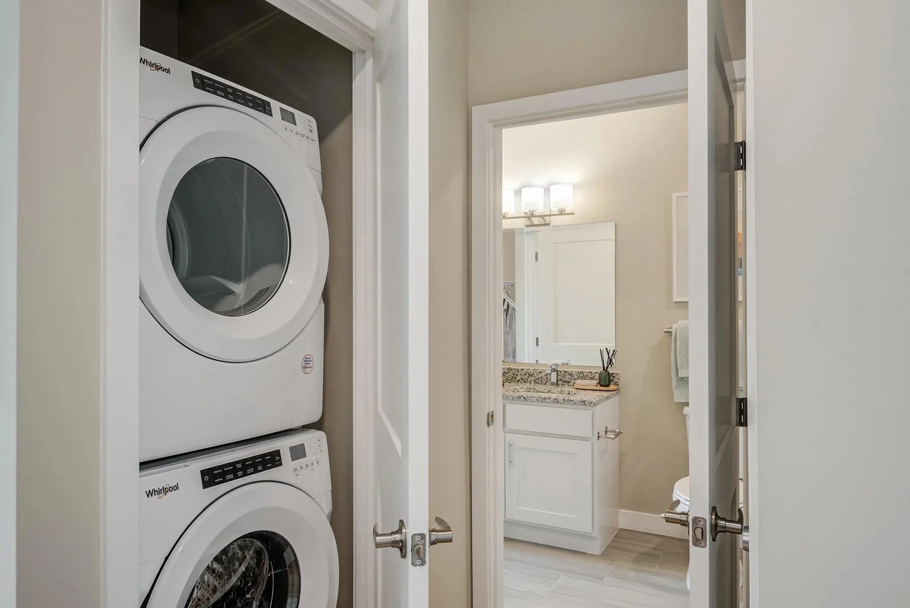 A laundry area with a stacked washer and dryer inside a closet, open door leading to a bathroom with a granite countertop and a mirror with lights above it.