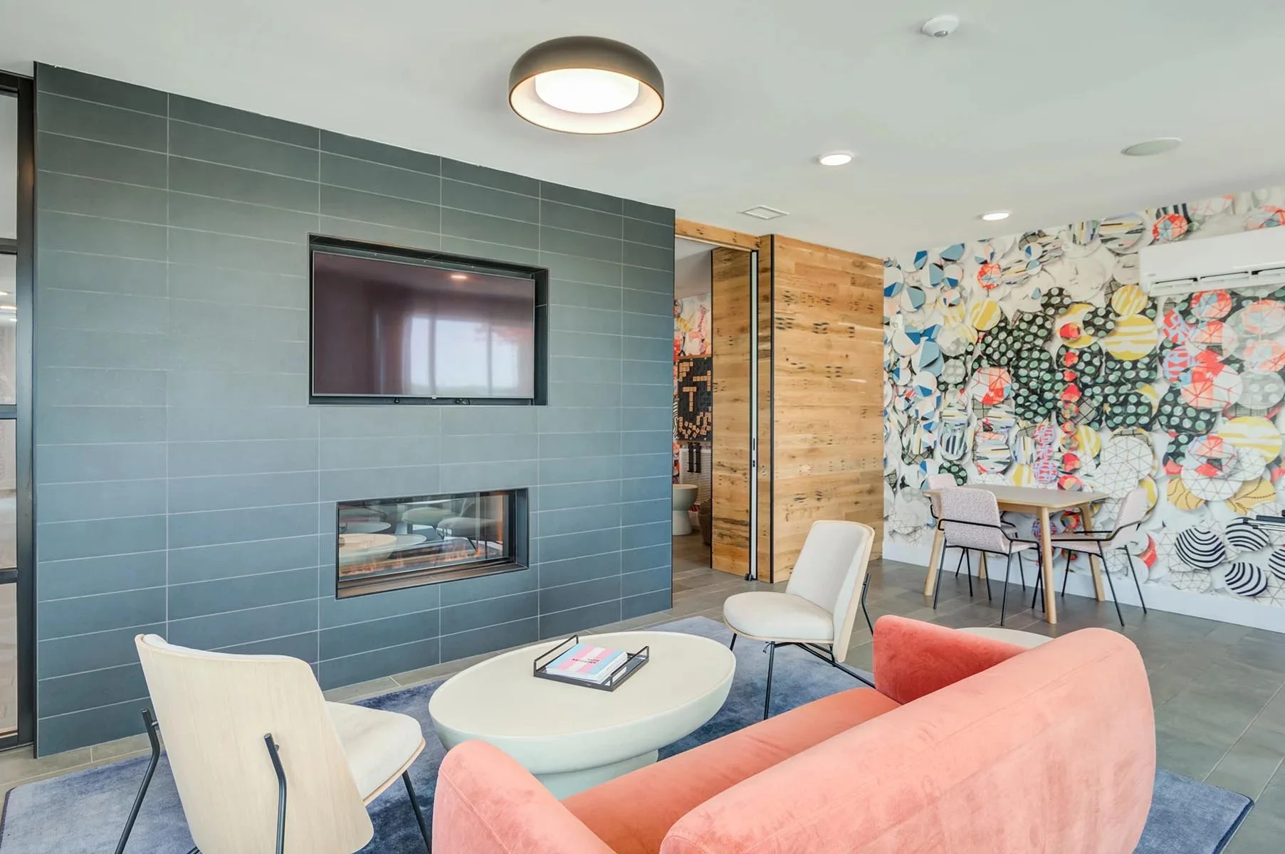 Modern room with gray tiled wall, wall-mounted TV, fireplace, colorful geometric patterned wallpaper, wooden partition, and contemporary furniture including plush peach sofa, white chairs, and a round coffee table with books.
