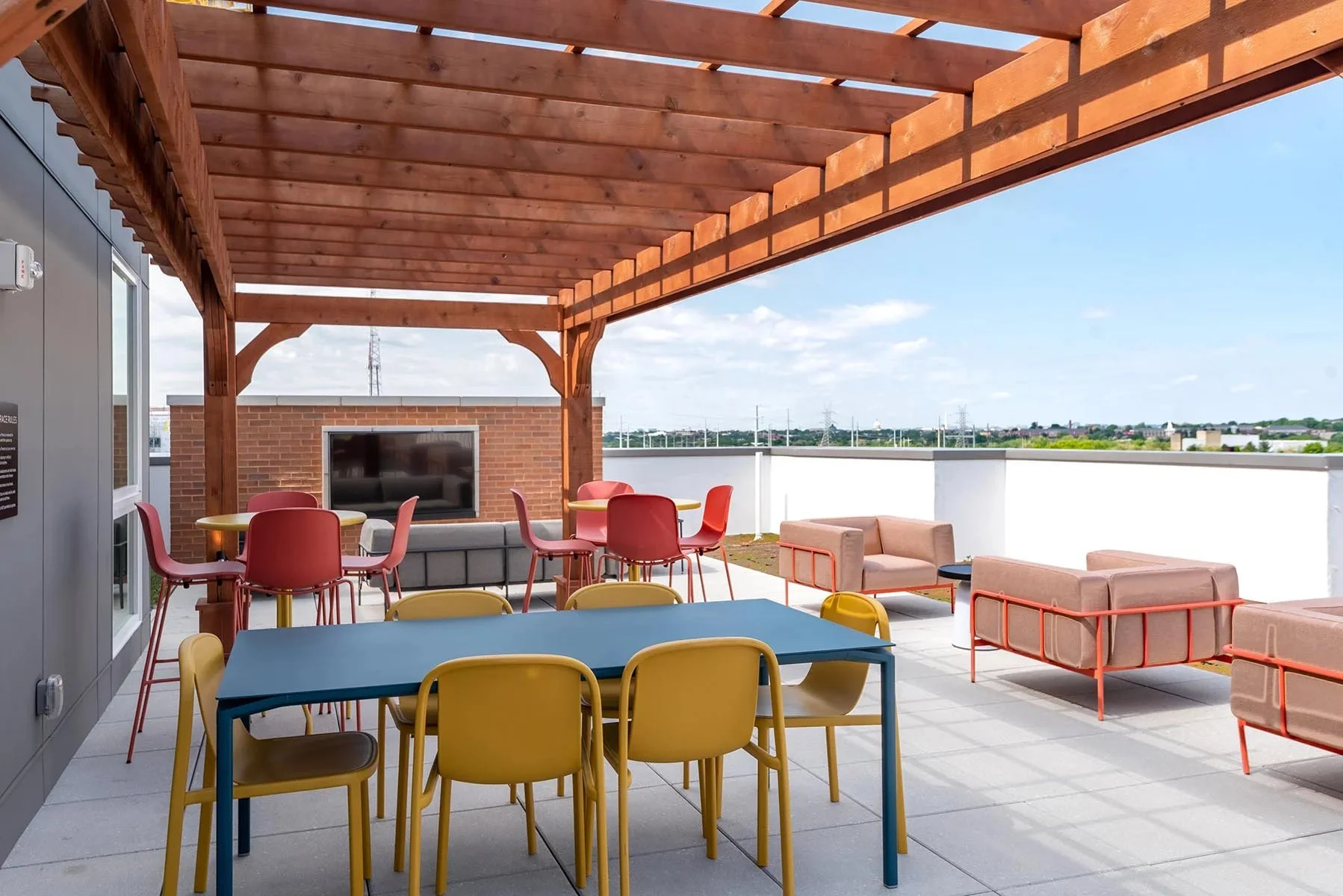 Rooftop patio with colorful chairs and tables, a wooden pergola overhead, and a sitting area with pink sofas, overlooking a cityscape and open sky.