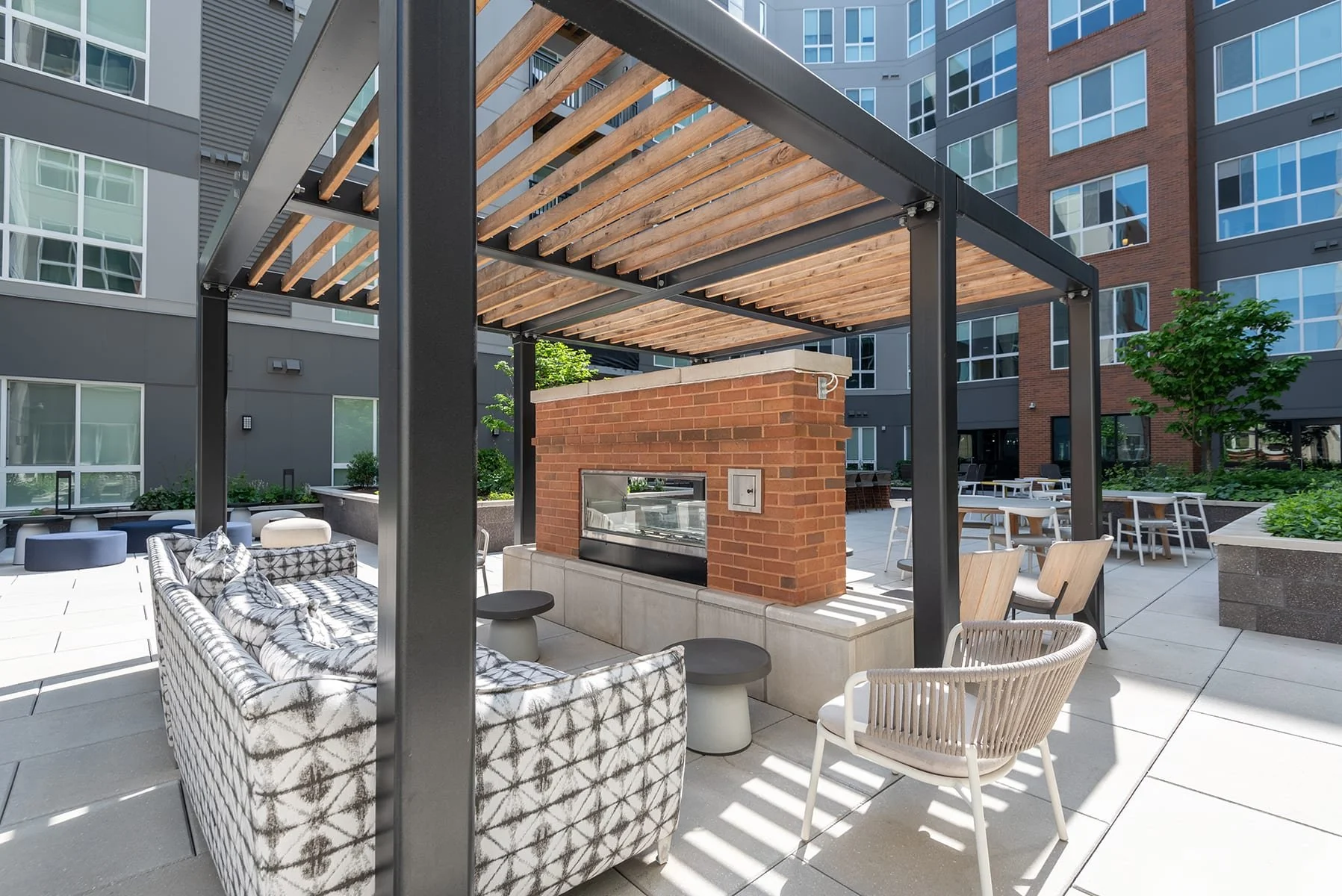 Outdoor patio area with modern furniture, a brick fireplace, and wooden canopy, surrounded by apartment buildings and landscaped greenery.