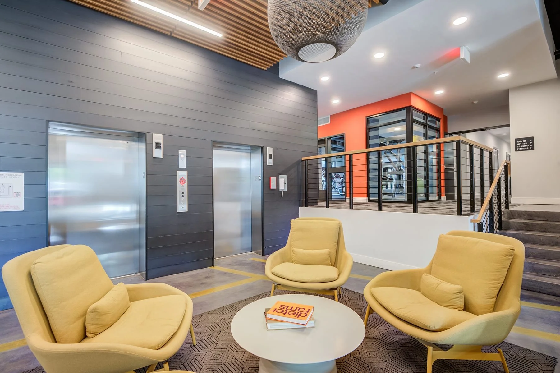 Modern lobby area with four yellow armchairs around a white round coffee table, on which there are books, one titled 'Ariel Sam'. Two elevator doors are in the background along with a staircase to the right, glass panels, and a brightly lit ceiling. 