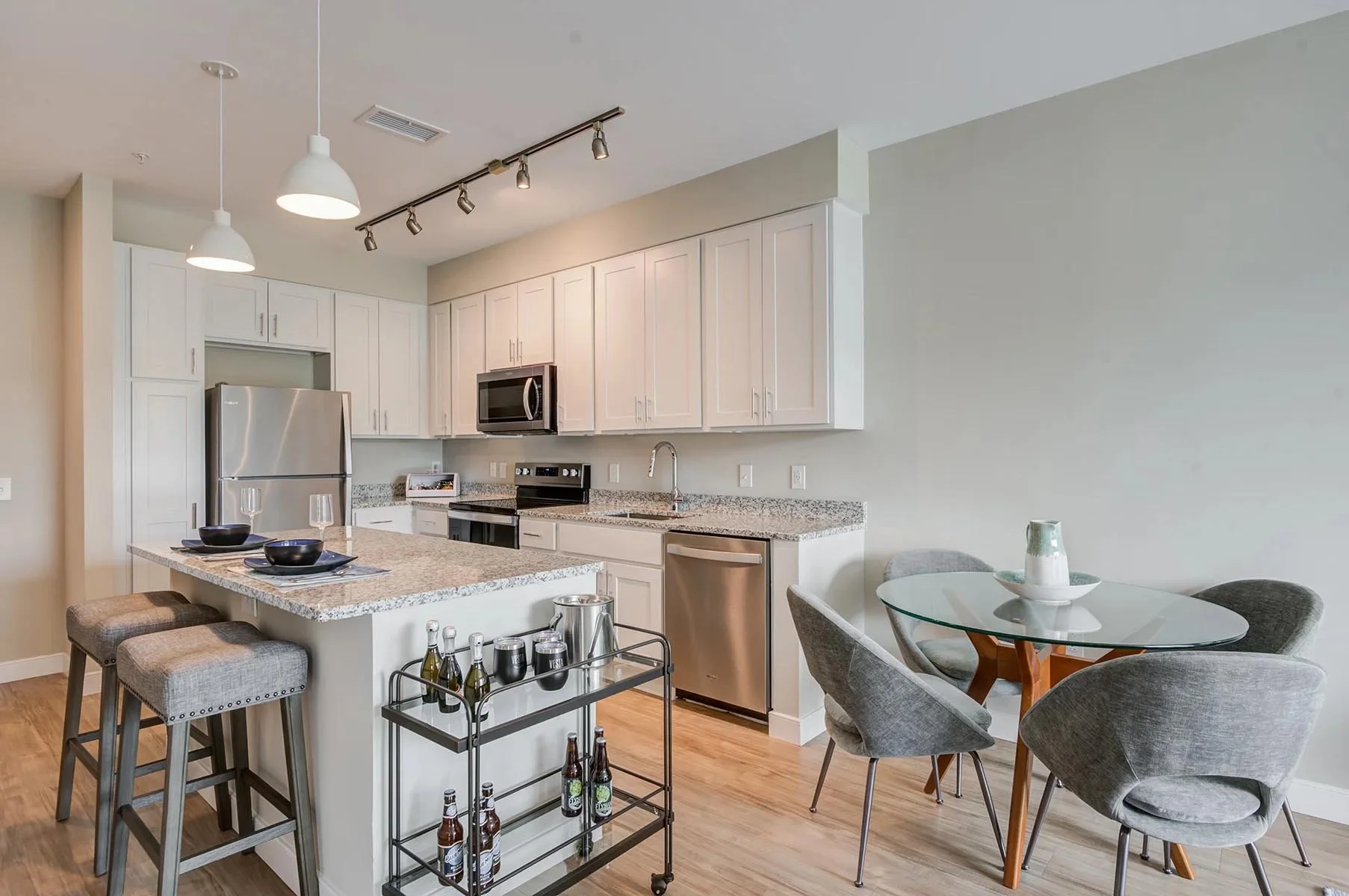Modern kitchen with white cabinets, stainless steel appliances, granite countertops, and a small dining area with four gray upholstered chairs around a glass table. An island with stools and a metal bar cart with drinks are also visible.