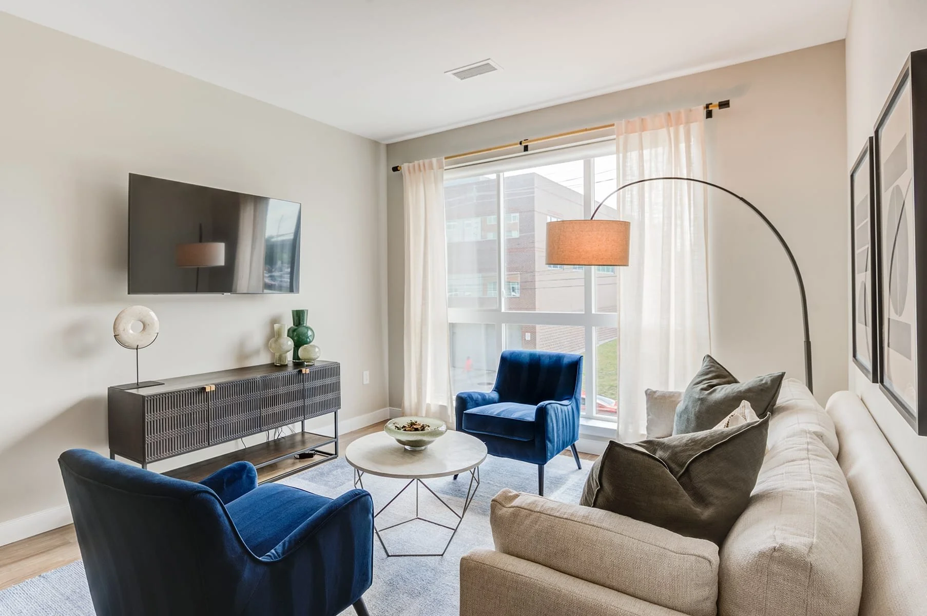 Living room with beige sofa, blue armchairs, coffee table, wall-mounted TV, and large window with curtains.