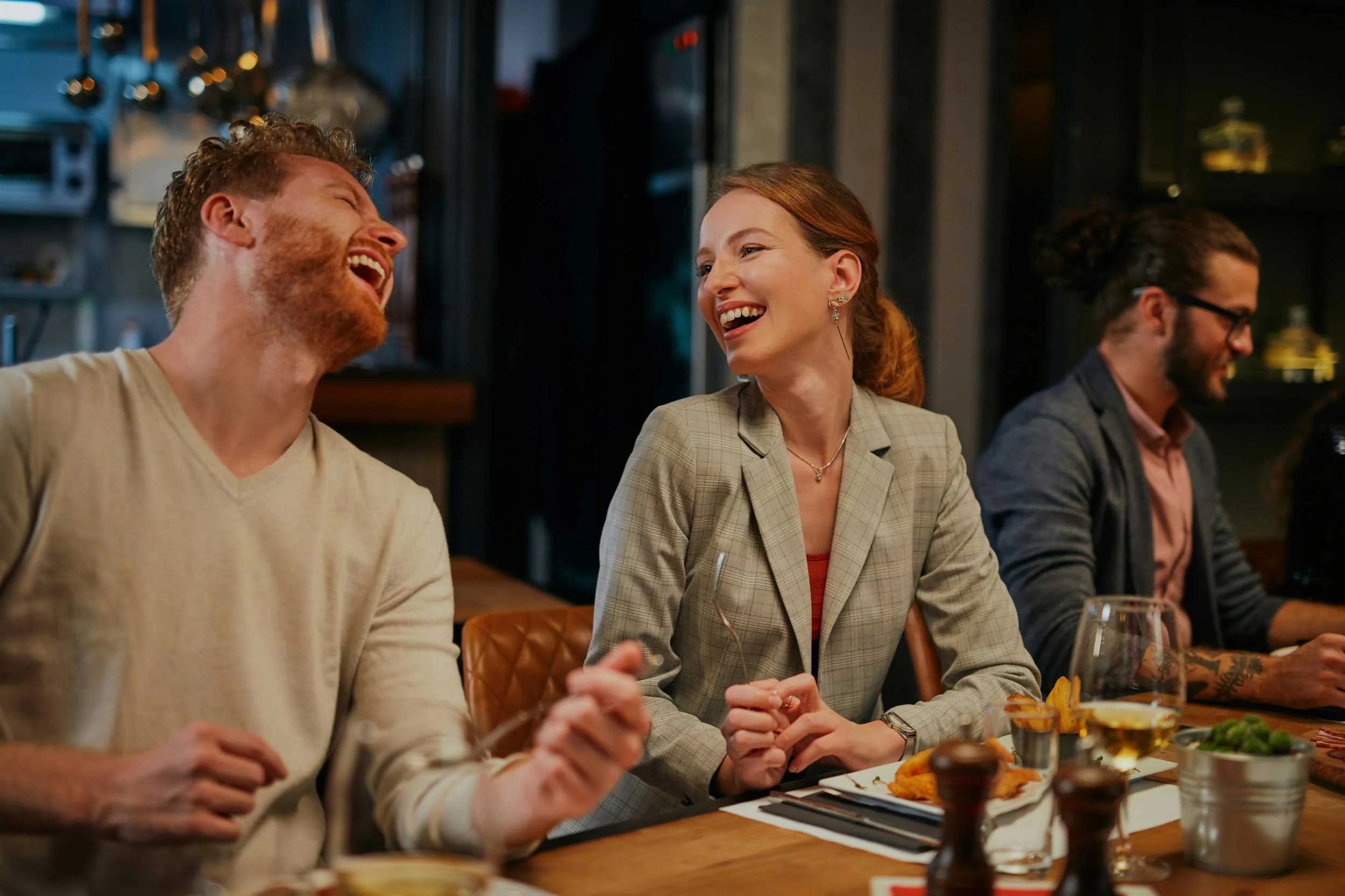 People sitting at a dinner table, laughing and smiling, with food and drinks in a cozy restaurant setting.