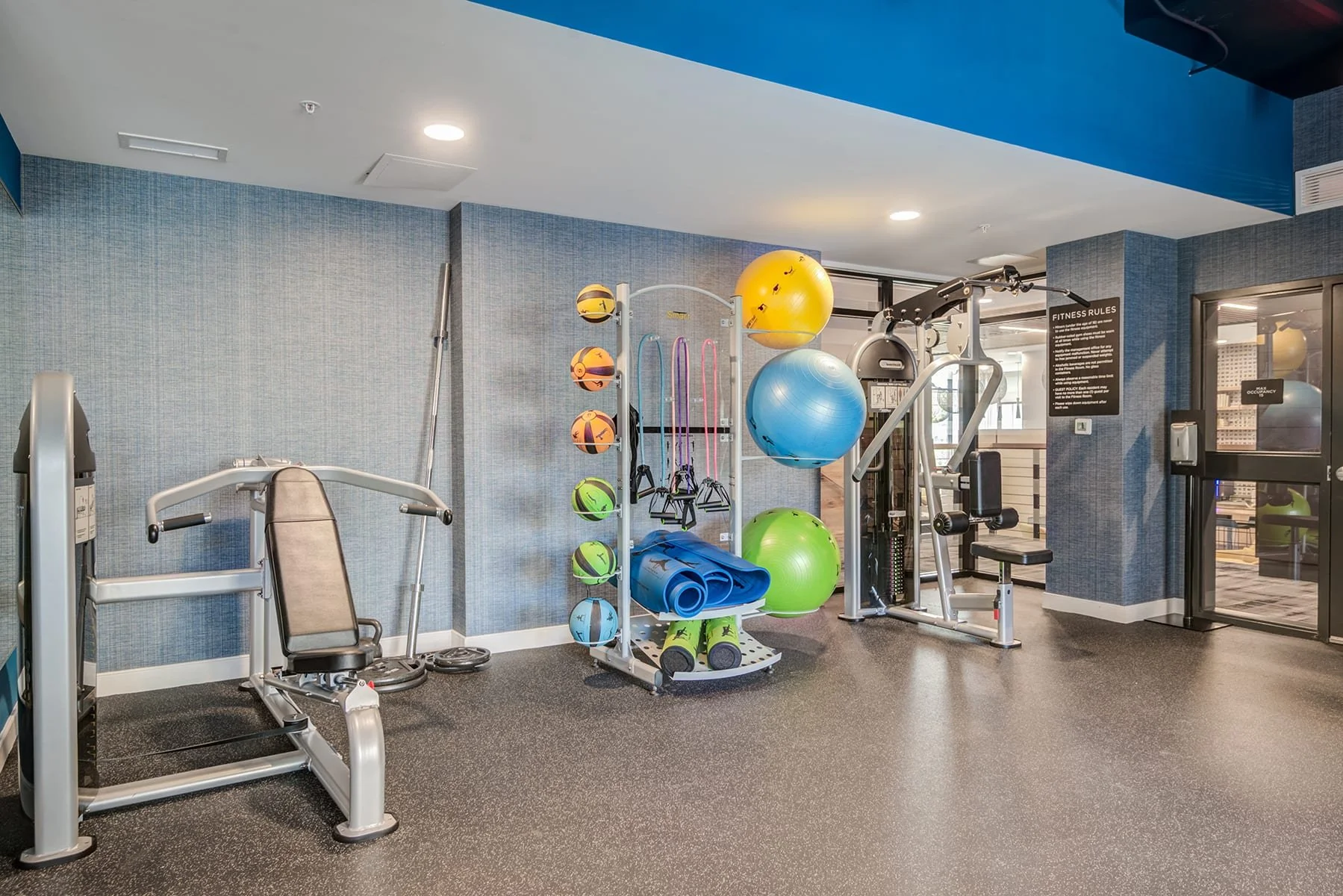 Fitness center with exercise equipment including stability balls, resistance bands, yoga mats, and a machine for strength training, with informational sign and lockable glass door.