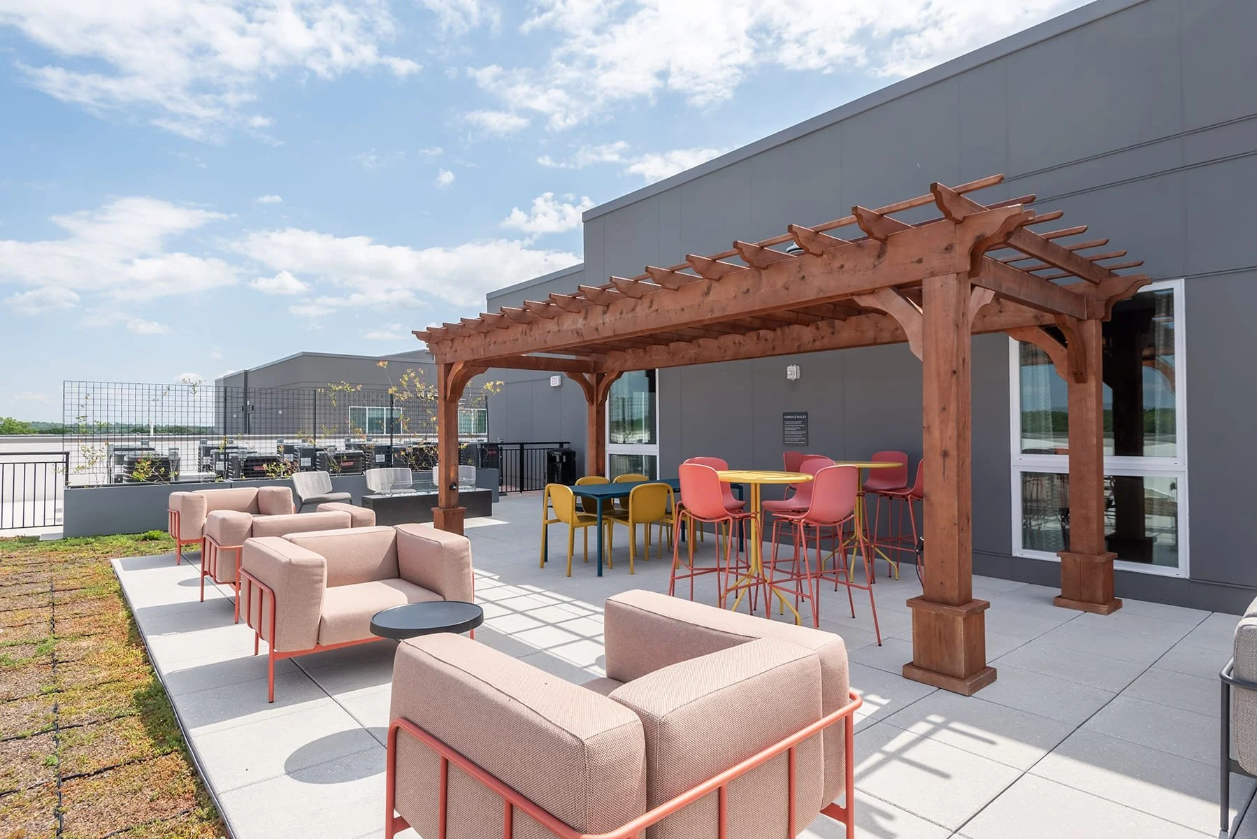 Outdoor rooftop lounge with pink cushioned seating, a wooden pergola, and colorful chairs around tables, with a modern building and blue sky in the background.