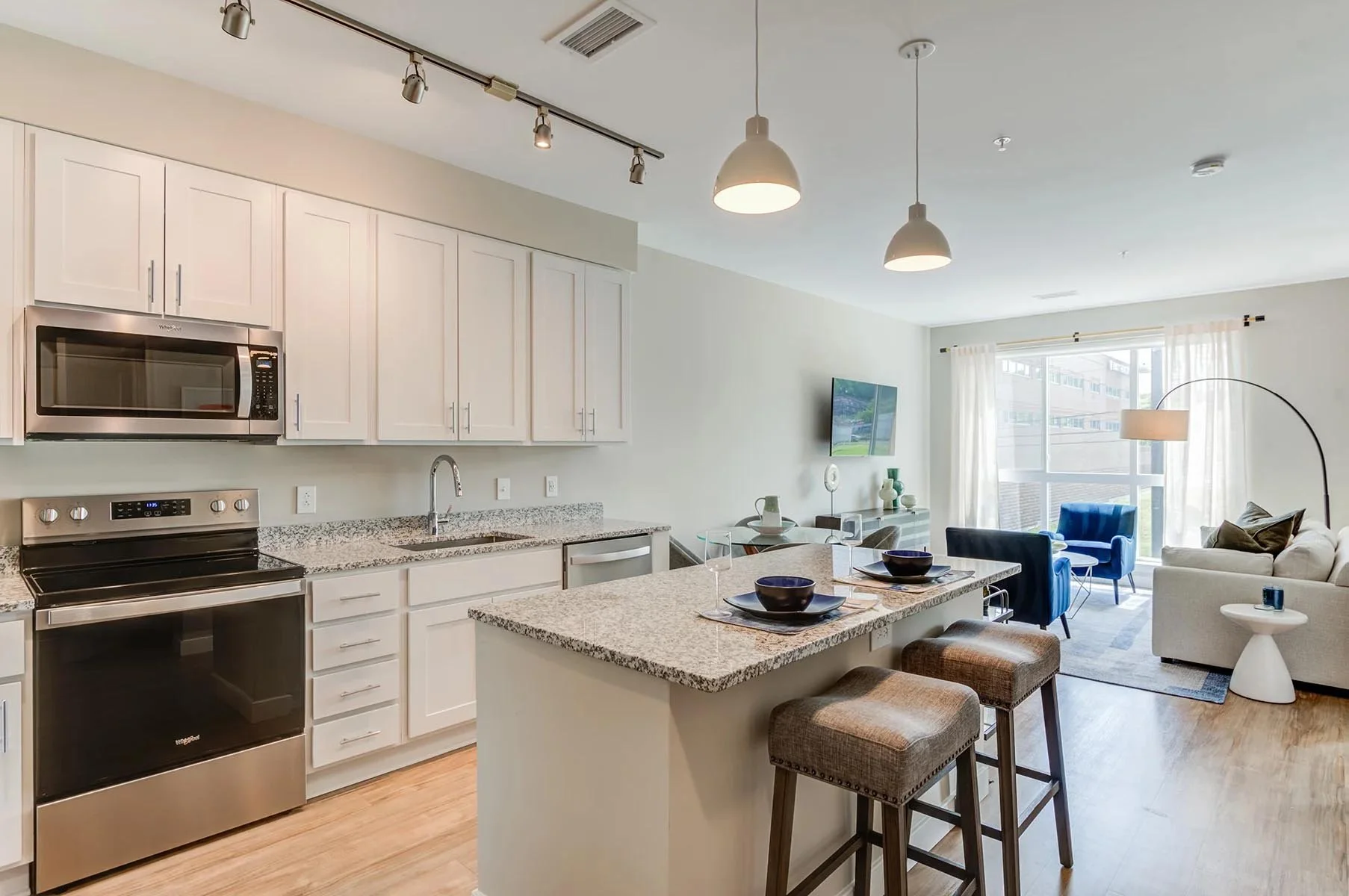 Modern kitchen with white cabinets, stainless steel appliances, granite countertop, and an open living area with large windows, blue and white furniture, and a flat-screen TV.