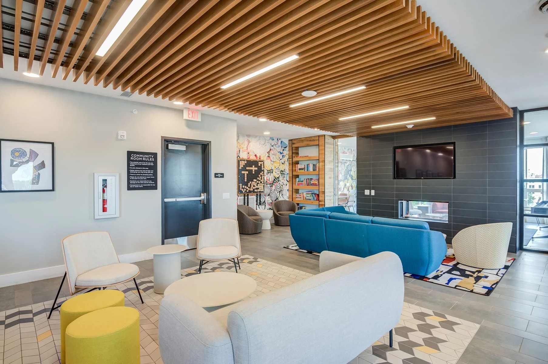 Modern lounge area with white, teal, and beige chairs and bright yellow ottomans, geometric patterned floor tiles, a fireplace with a television above, and a wooden ceiling feature with lights.