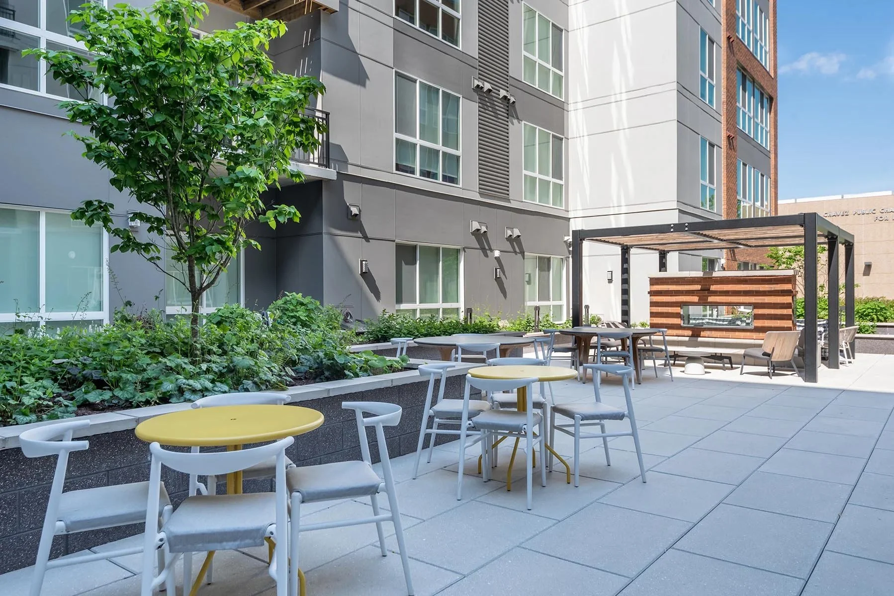 An outdoor patio area with yellow and white tables and chairs, surrounded by green plants and a small tree, with a modern multi-story apartment building in the background under a clear blue sky.