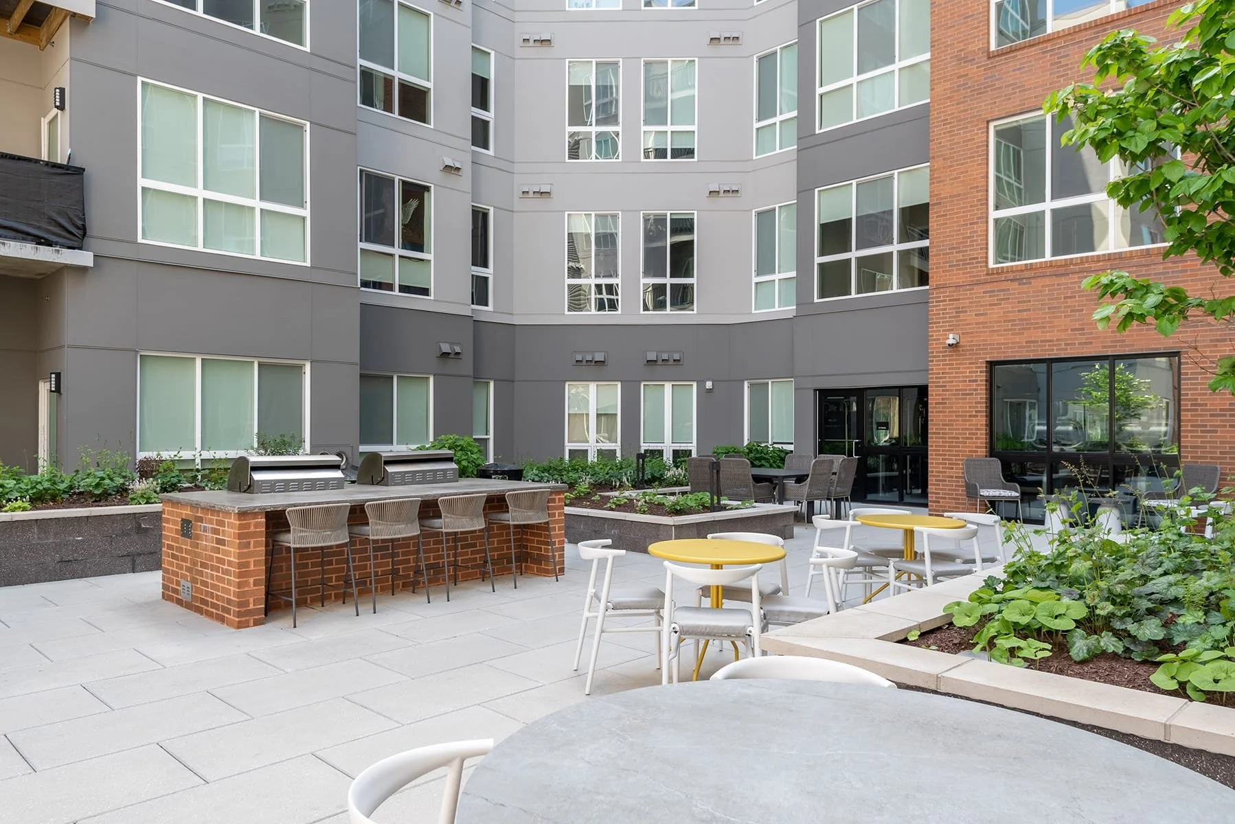 A modern outdoor patio with white and yellow tables and chairs, surrounded by green plants, with a gray and brick apartment building in the background.