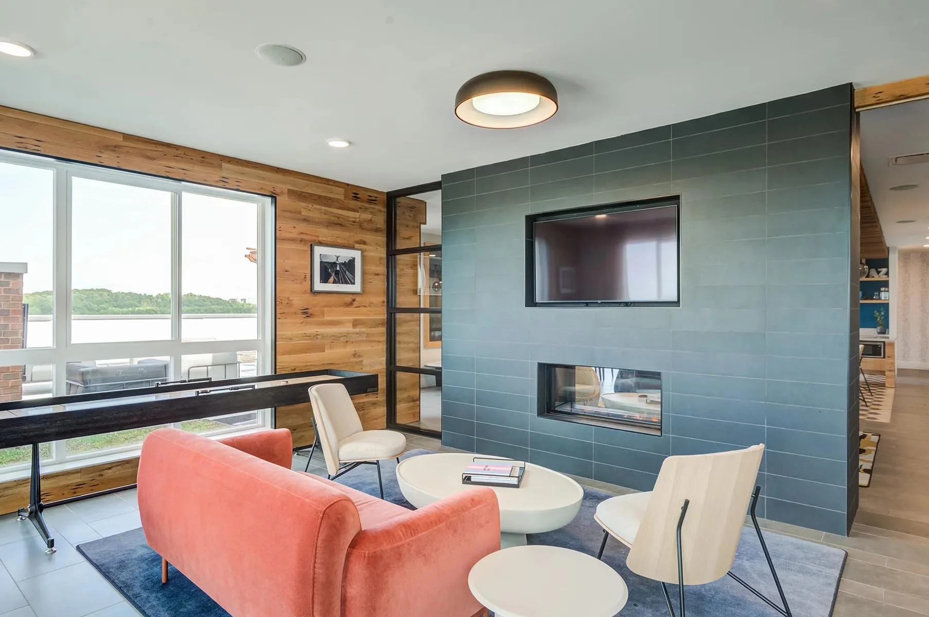 Community room with large window, wooden wall, blue tiled fireplace with a TV, coral sofa, white chairs, coffee table, and a view of a partly cloudy sky and trees outdoors.