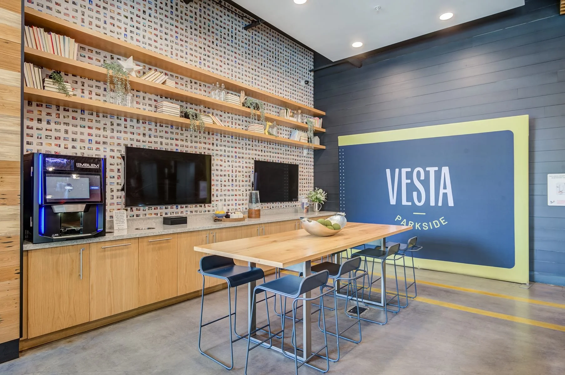 A modern indoor lounge or communal area with a wooden table, chairs, a large sign that reads 'VESTA PARKSIDE,' a kitchenette with a coffee machine, and decorative shelves with books and plants.