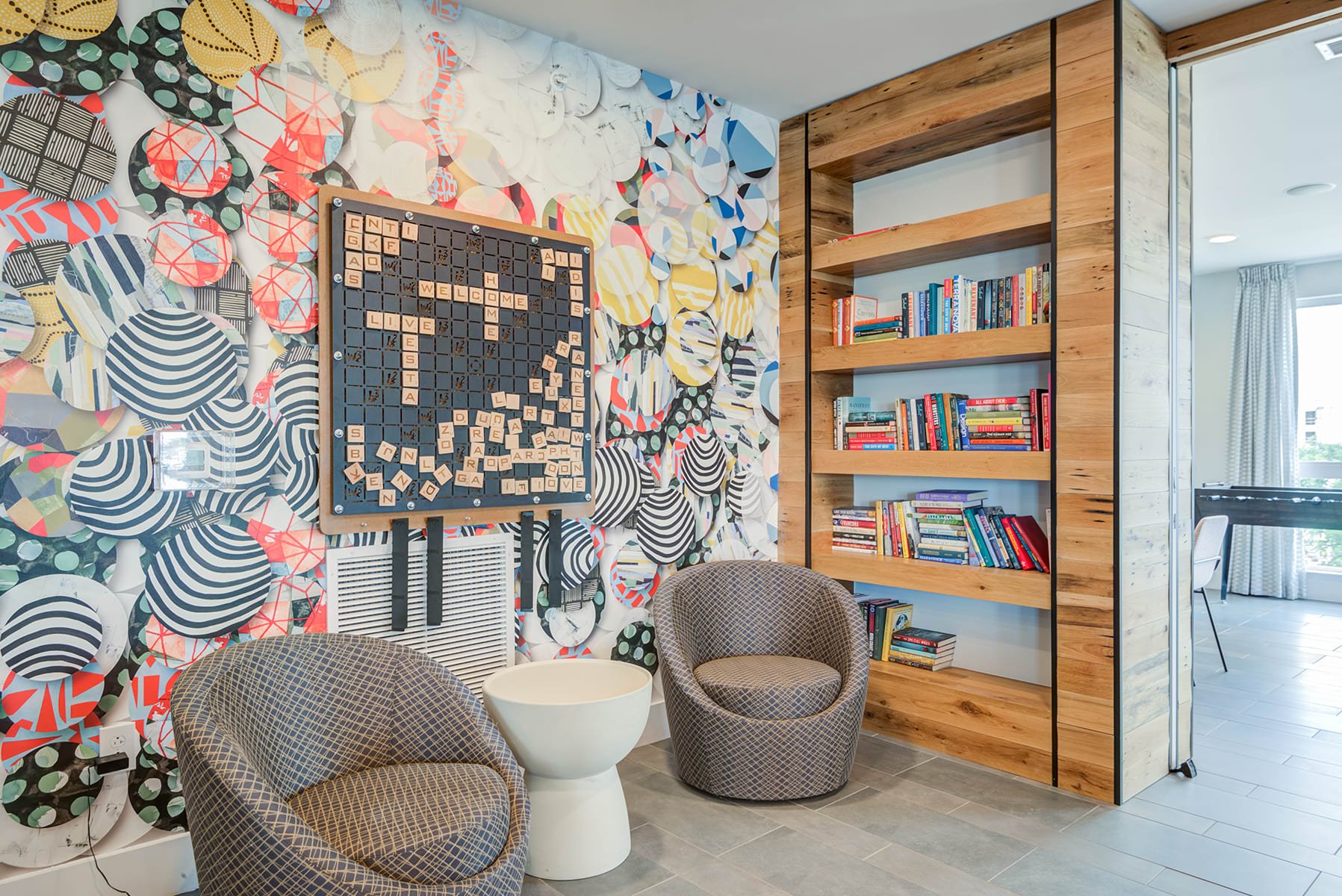 A room with a colorful, abstract-patterned wall, a black scrabble board with words, a wooden bookshelf filled with books, and seating including two patterned chairs and a small white table.