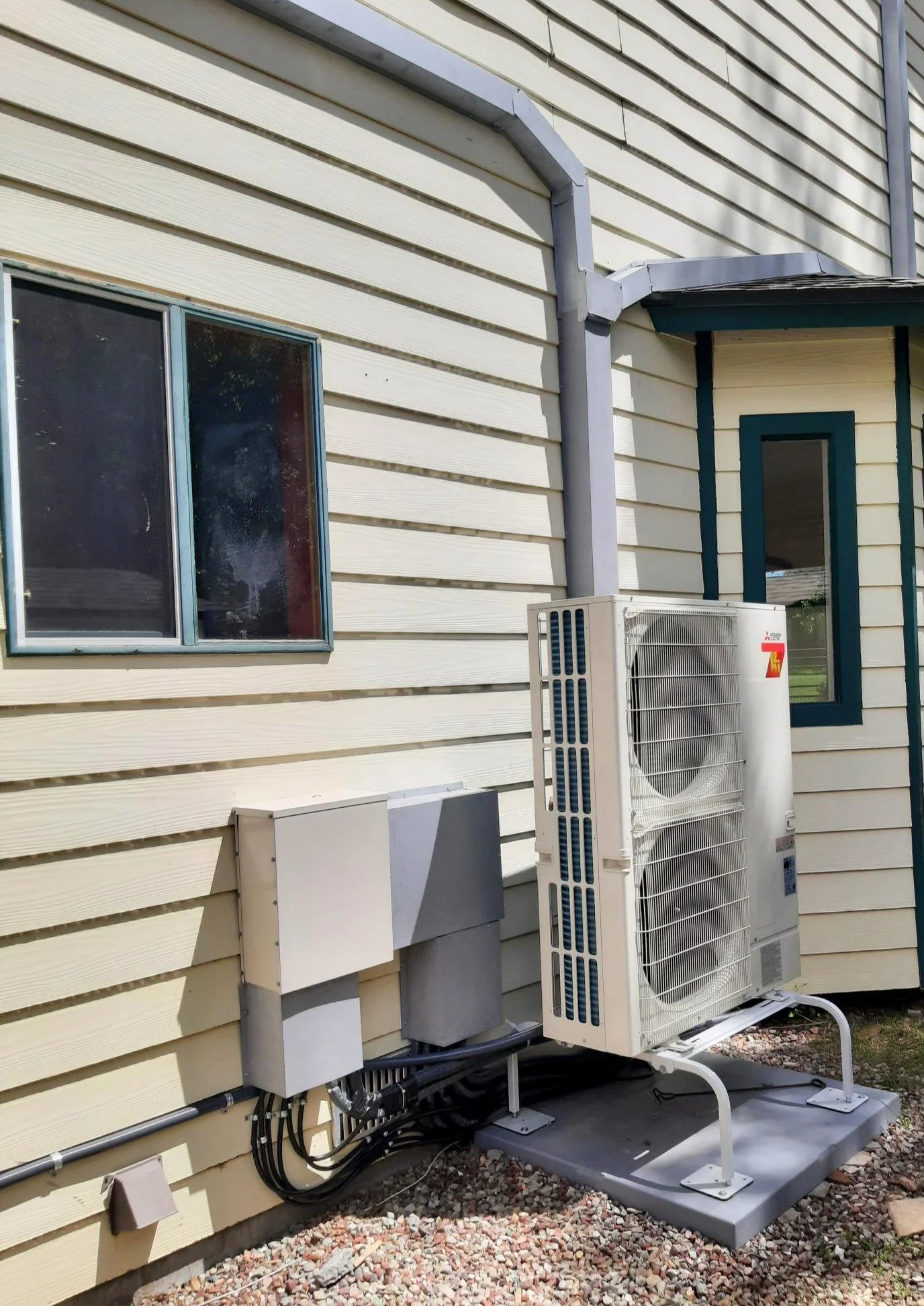 An outdoor view of a house with yellow siding, two windows, and an air conditioning unit installed on the ground with black cables connected to it.
