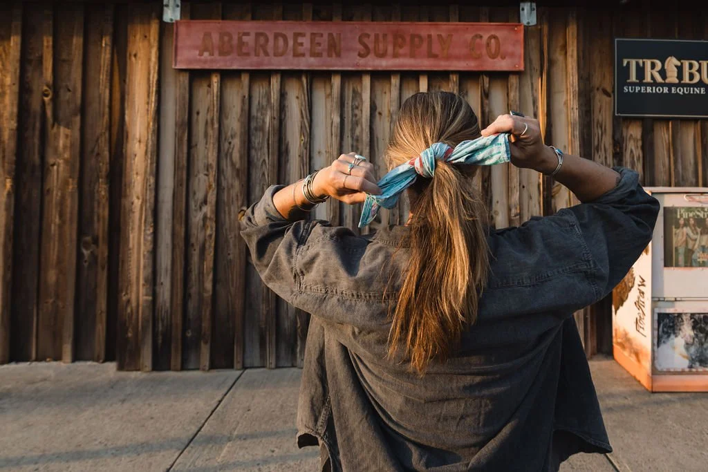 A woman with long hair tying a colorful scarf around her head in front of a wooden wall with signs, one reading 'ABERDEEN SUPPLY CO.' and another for a horse-related business.