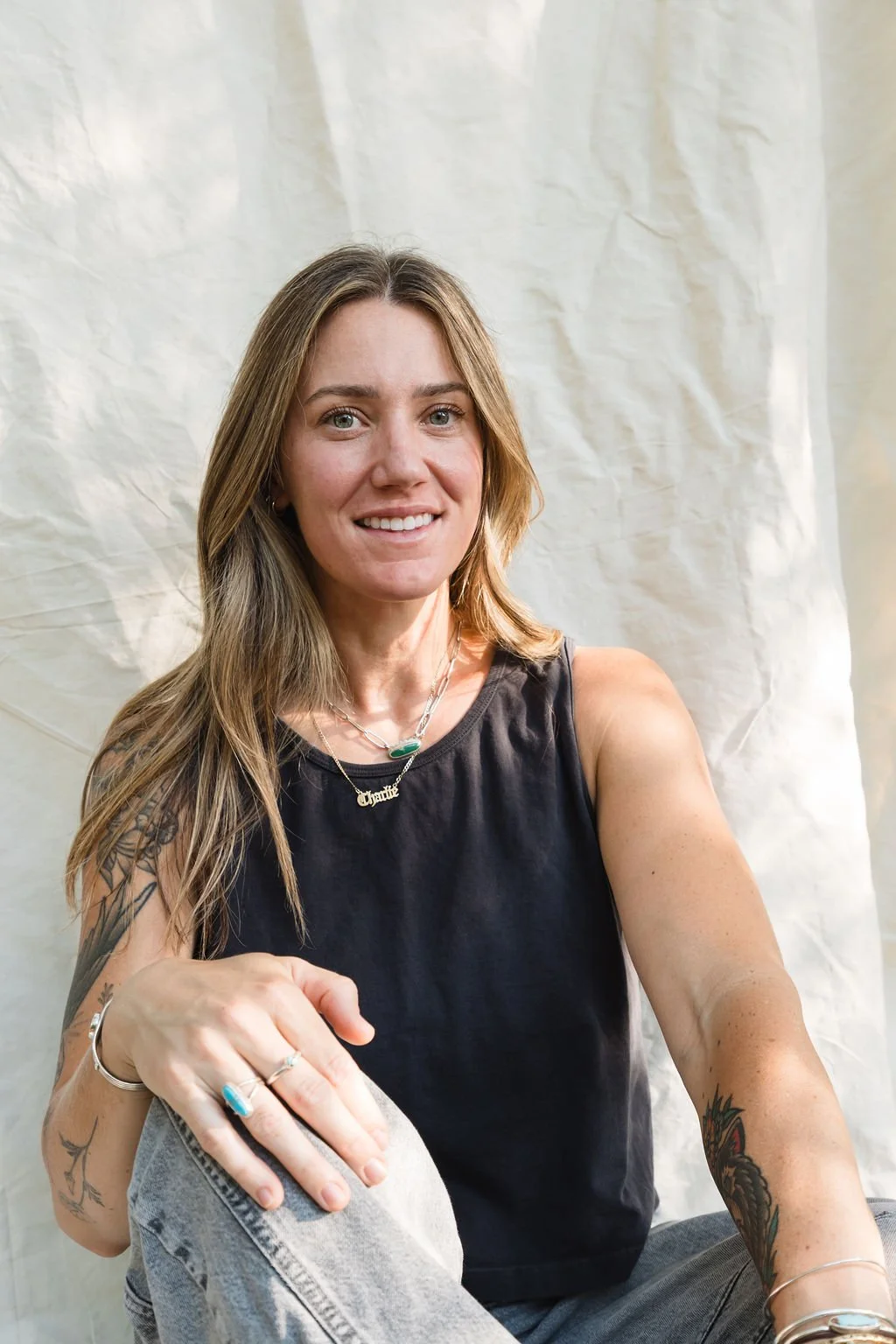 Tommor Sun Founder, Allie wearing a black sleeveless top and jewelry, sitting against a plain light-colored background and smiling at the camera.