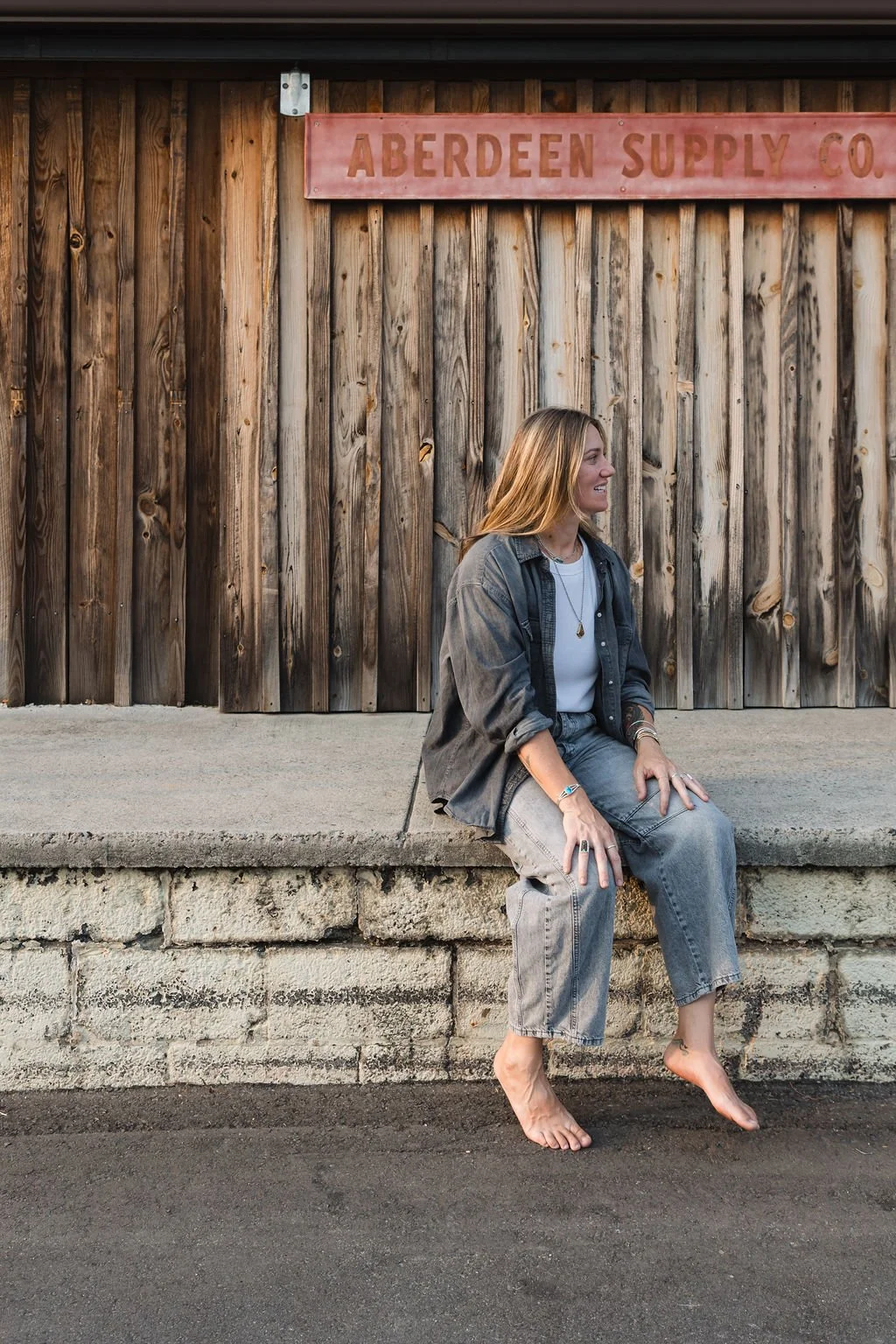 A woman with blonde hair sitting barefoot on a concrete ledge in front of a wooden wall with a pink sign reading 'Aberdeen Supply Co.' She is smiling and looking to her right, dressed in casual gray jeans, a white t-shirt, and a gray jacket, with jewelry on her hands and neck.