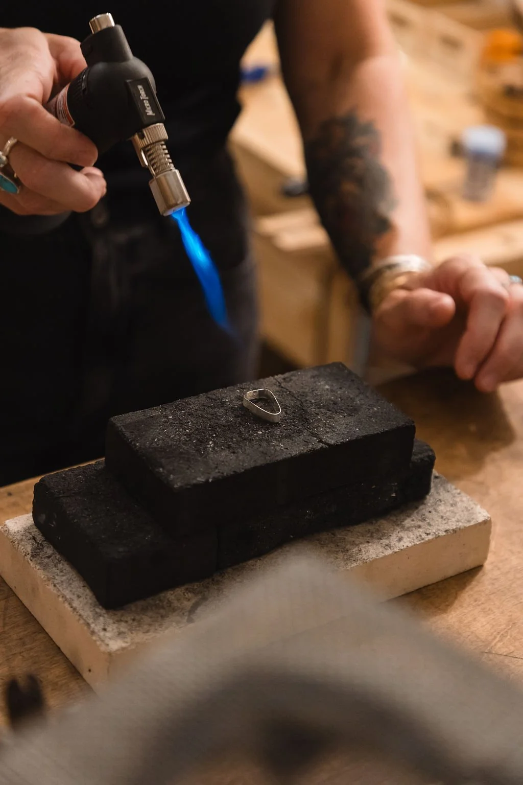 A person is using a blowtorch to work on a small metal ring placed on a black firebrick, on a wooden workbench.