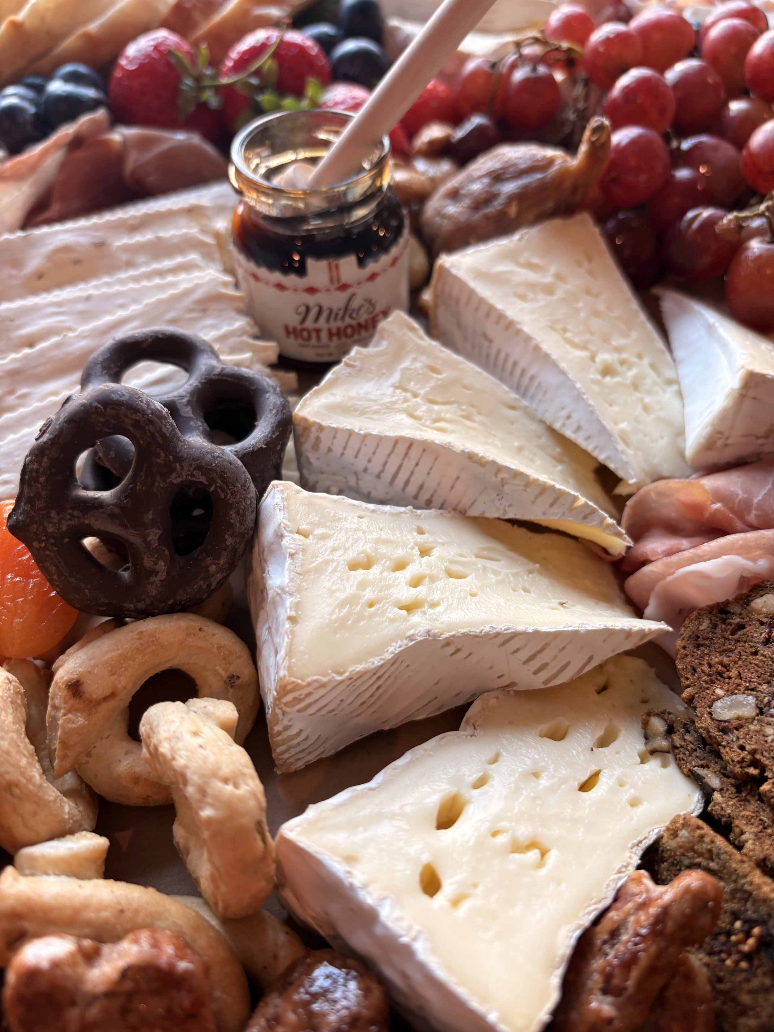 Close-up of a cheese platter with various cheeses, crackers, grapes, strawberries, a jar of honey, and other snacks.