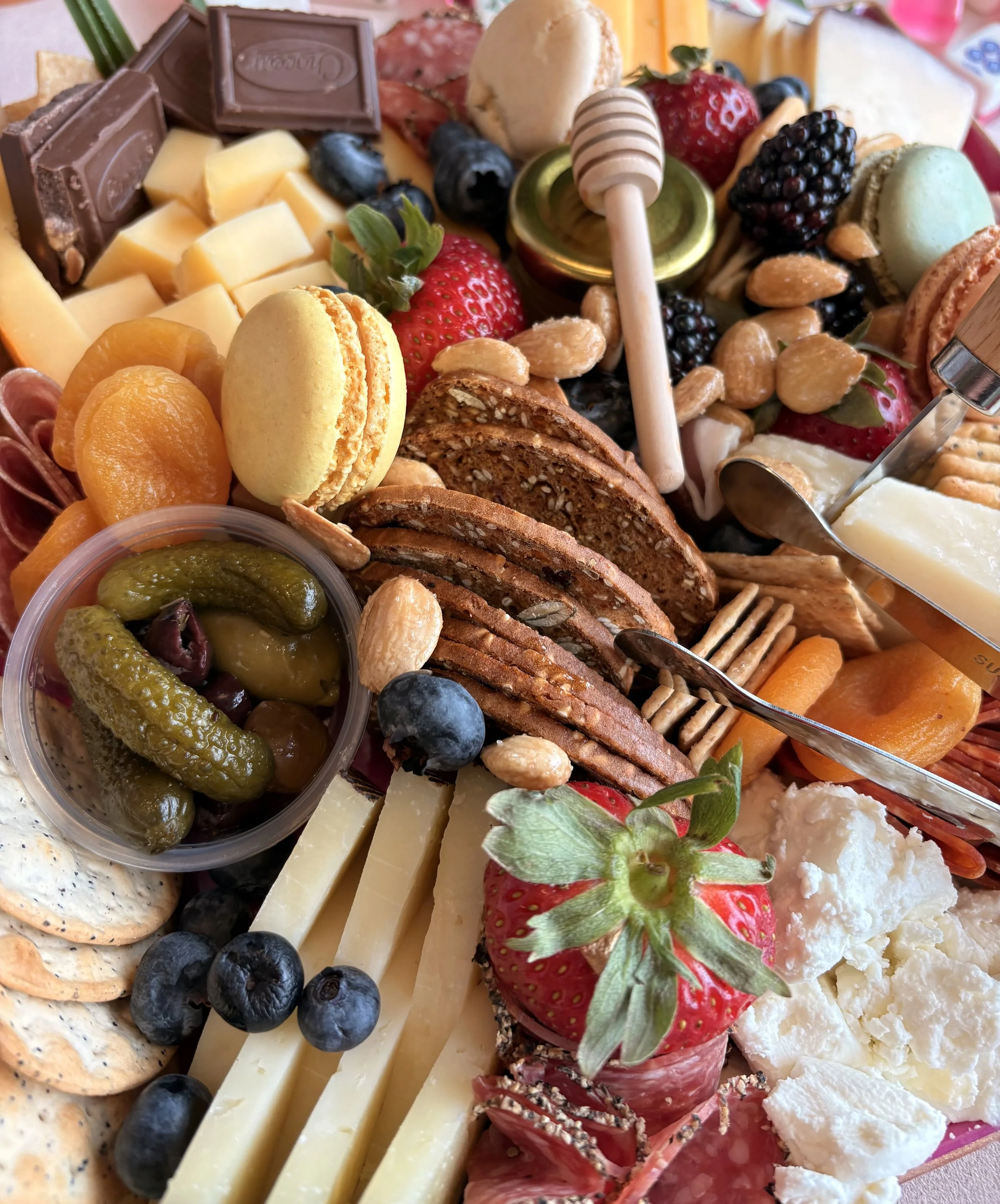 A platter of assorted foods including cheese, strawberries, blueberries, blackberries, almonds, crackers, chocolate, macarons, and pickles.