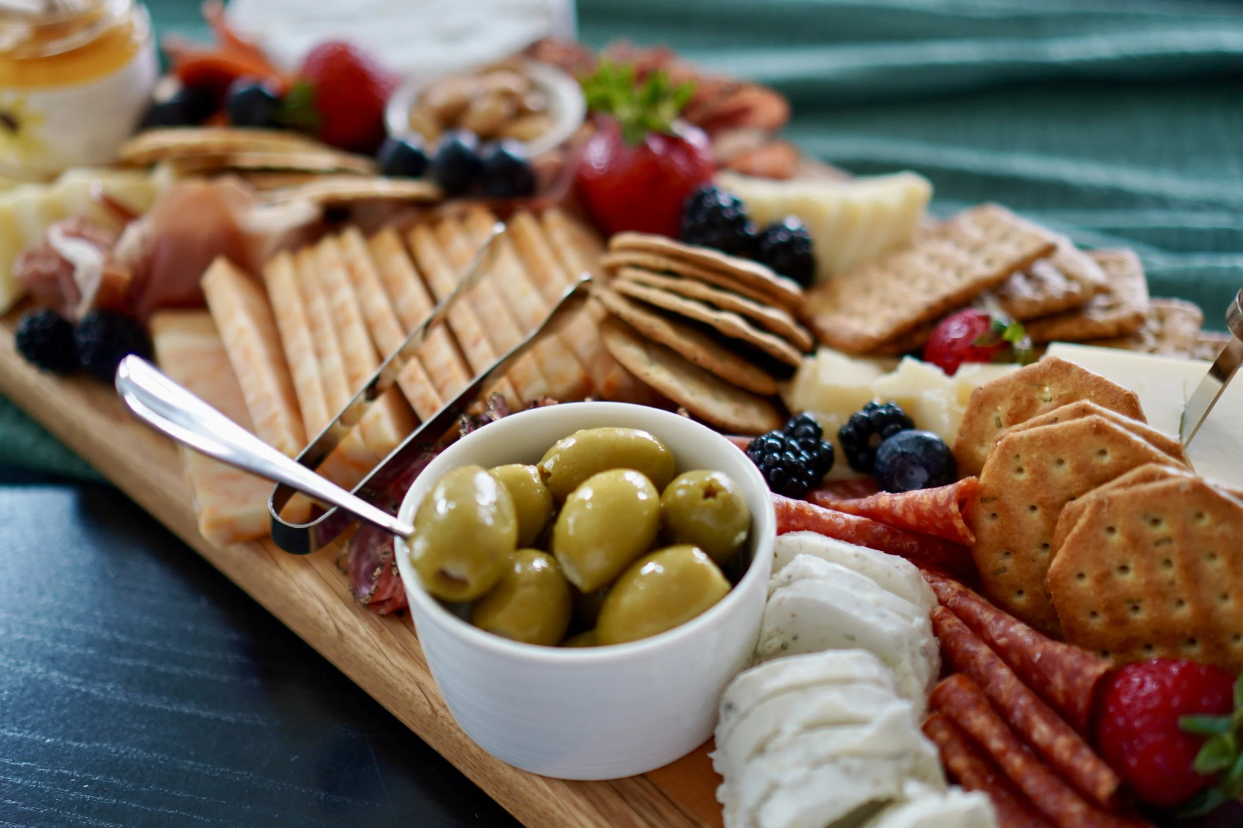A wooden platter with assorted cheese, crackers, green olives, and fresh berries on a dark table.
